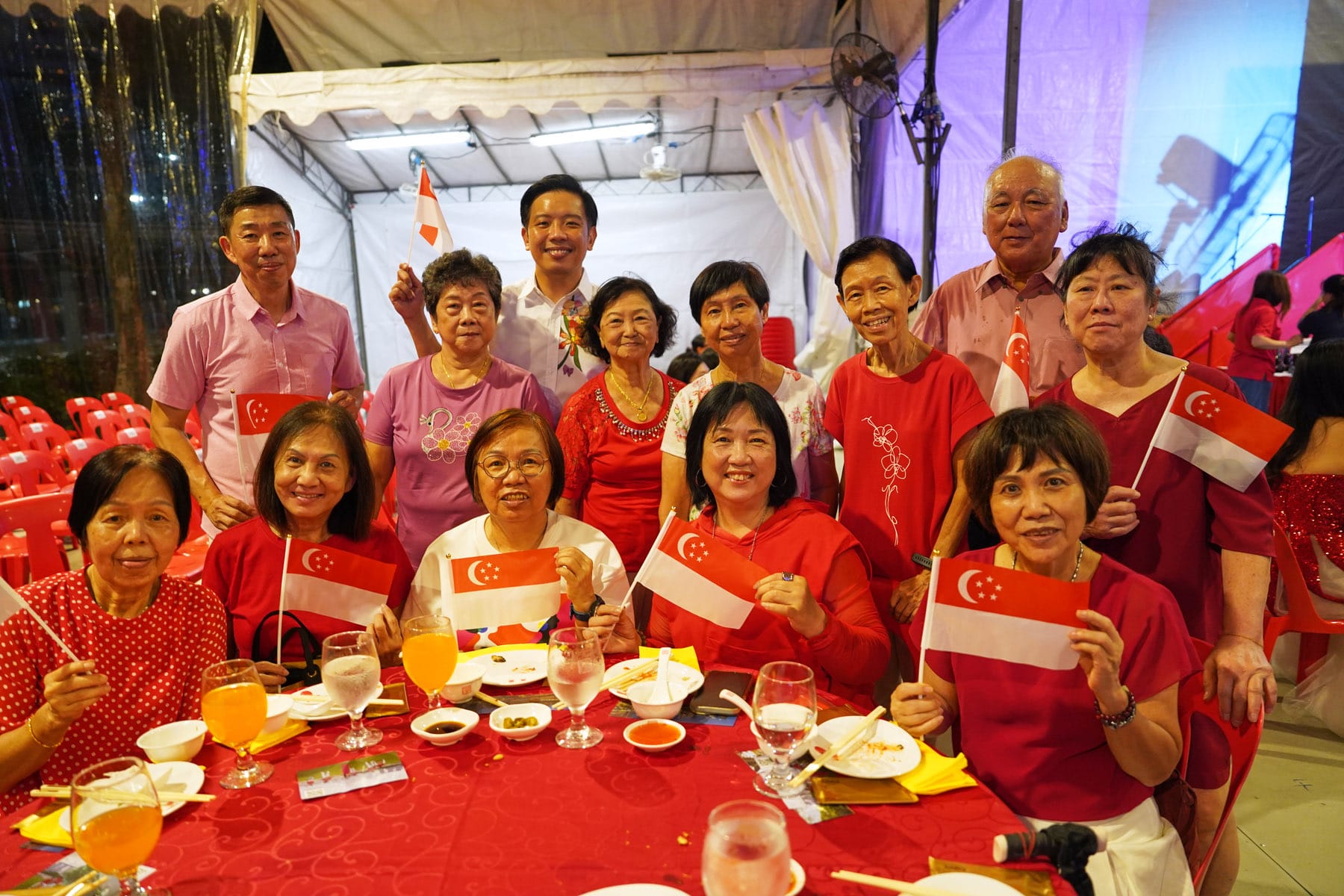 A group of smiling people in red clothing hold Singapore flags at a festive dinner table outdoors at night.