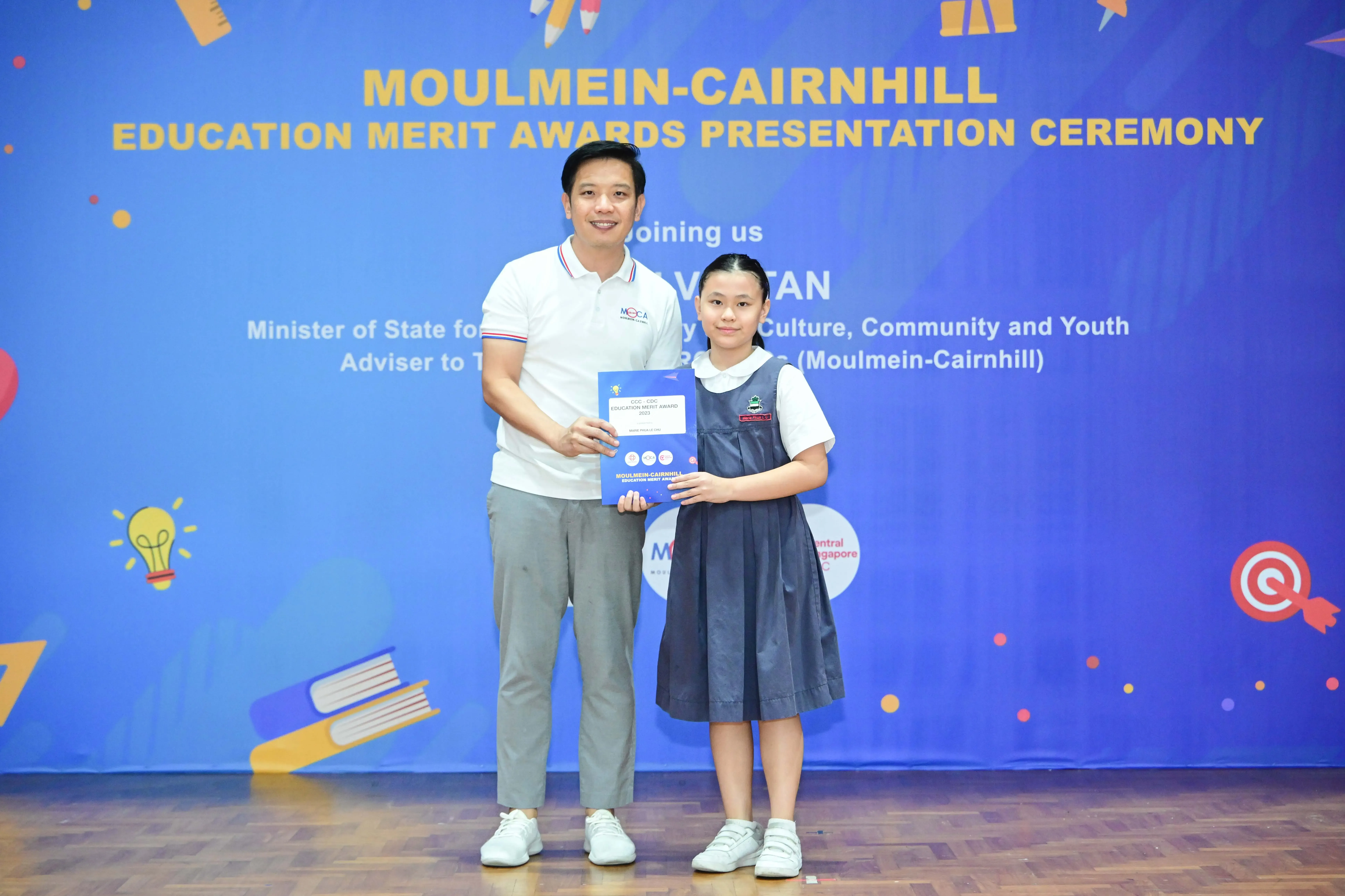 A man and a young girl in school uniform hold an award certificate at the Moulmein-Cairnhill Education Merit Awards ceremony.