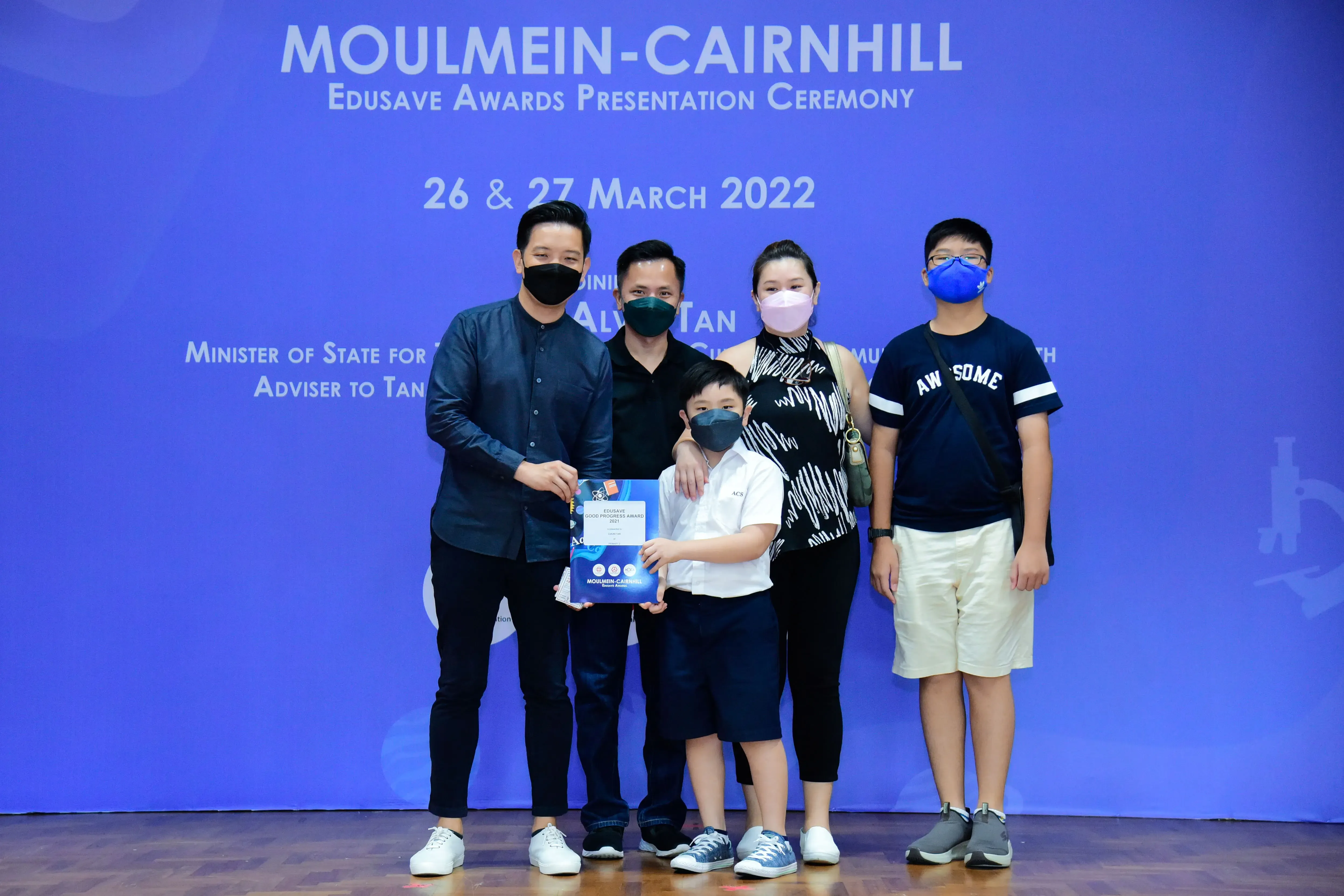 Five masked people pose at the Moulmein-Cairnhill Edusave Awards Ceremony on 26-27 March 2022, holding a certificate.