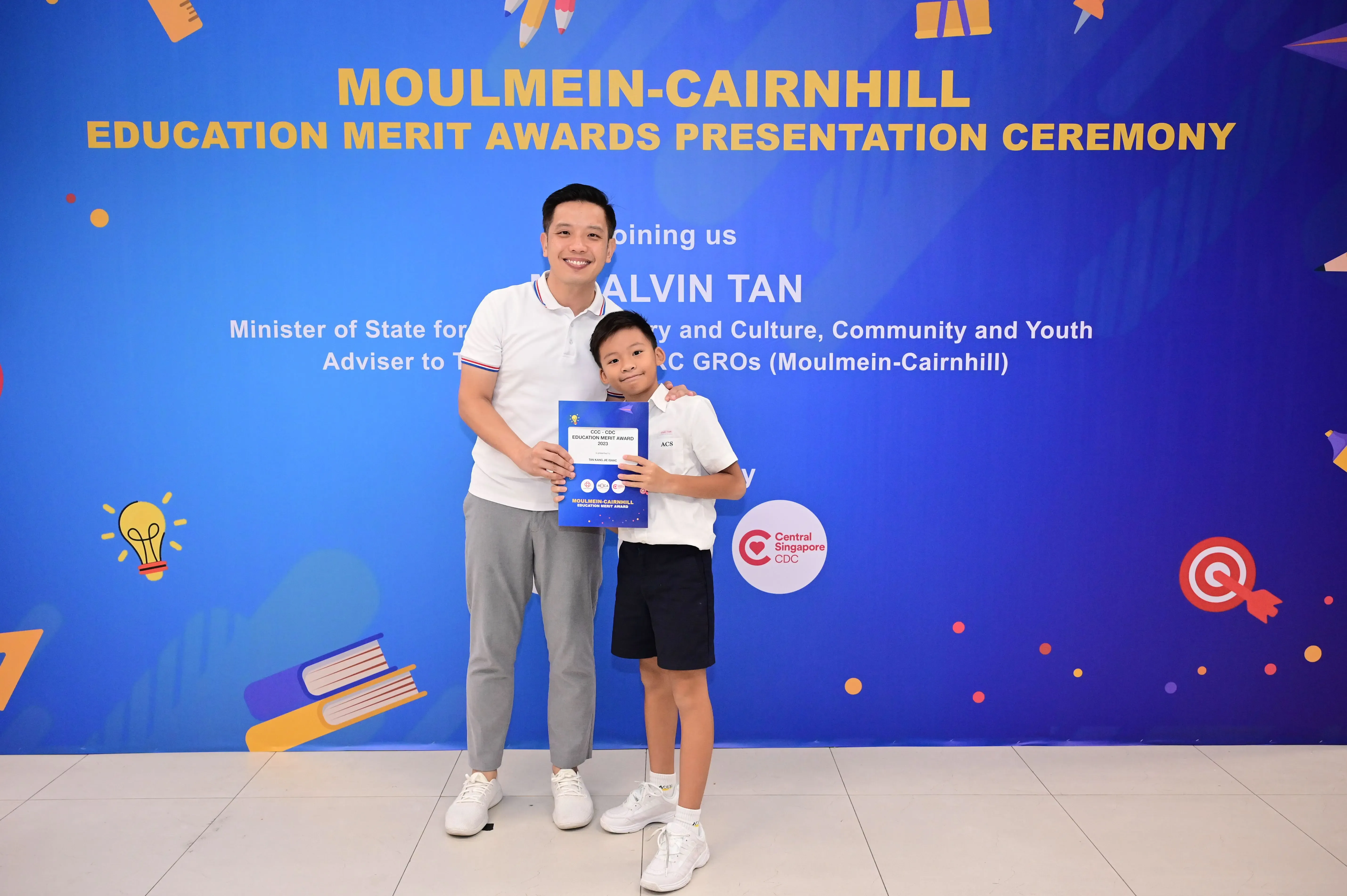 A man and a young boy pose together holding a merit award certificate in front of a blue education ceremony banner.