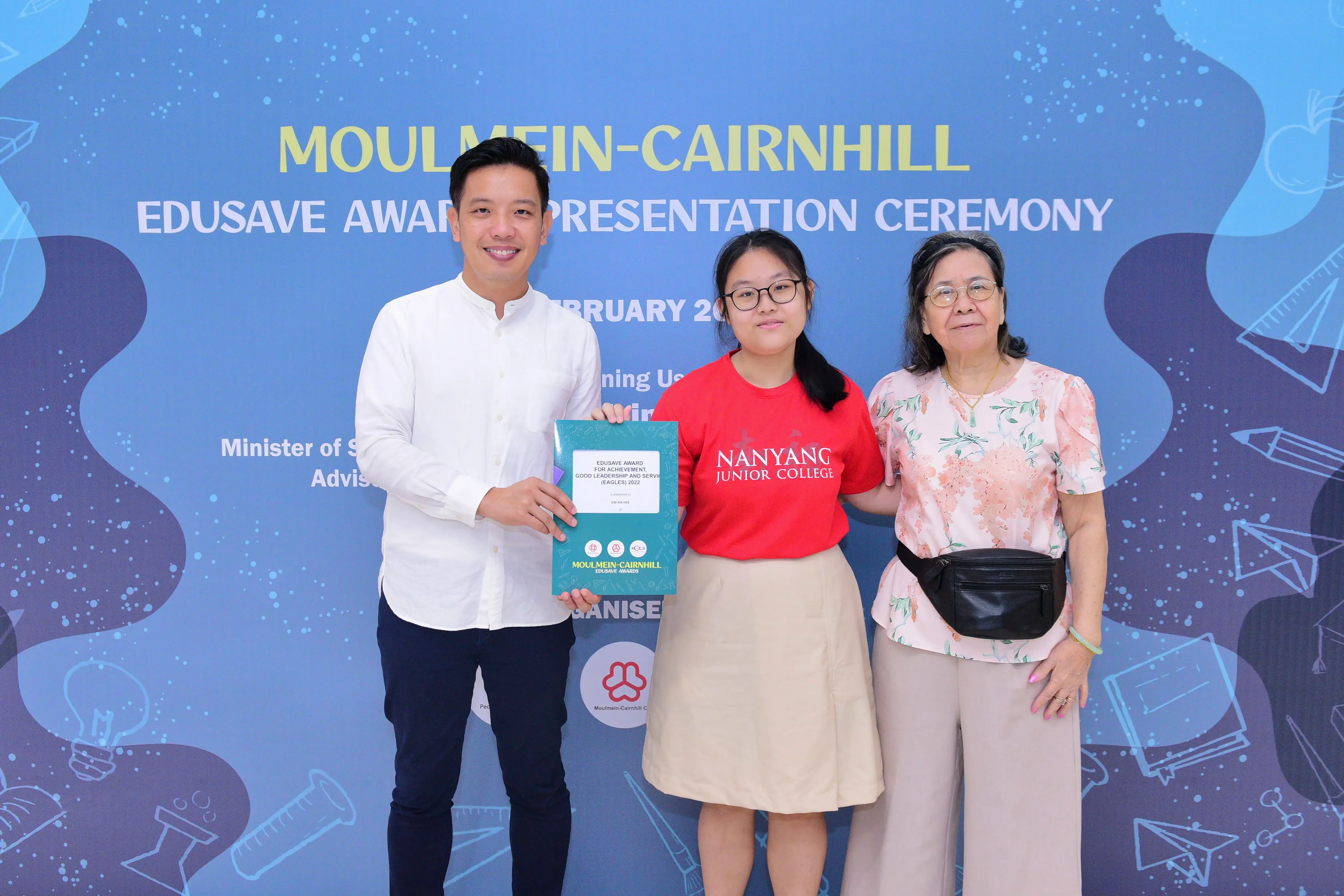 Three people pose holding an award booklet at the Moulmein-Cairnhill Edusave Awards Presentation Ceremony backdrop.