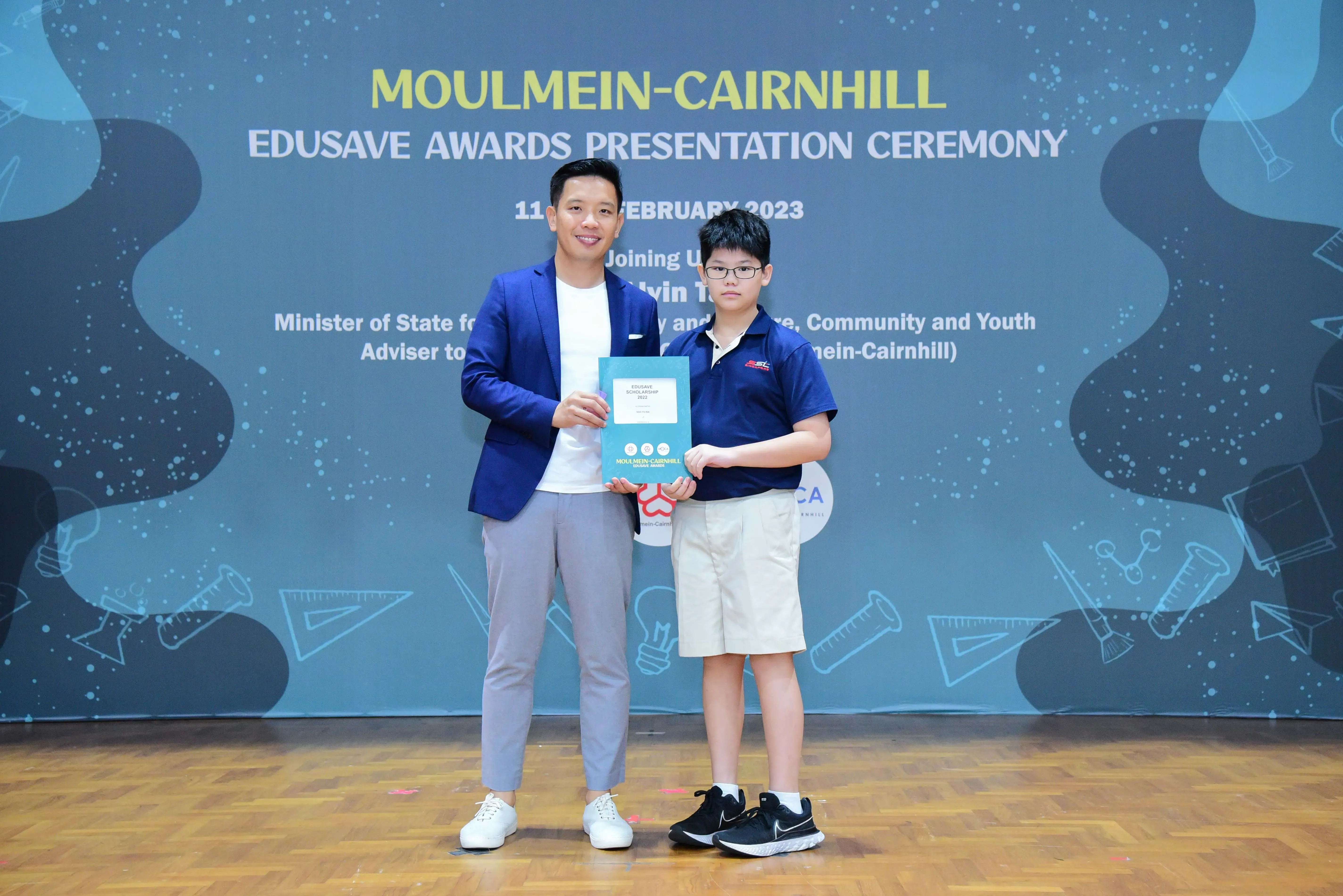 An adult in a blue blazer and a young boy hold a certificate at the Moulmein-Cairnhill Edusave Awards Ceremony 2023.