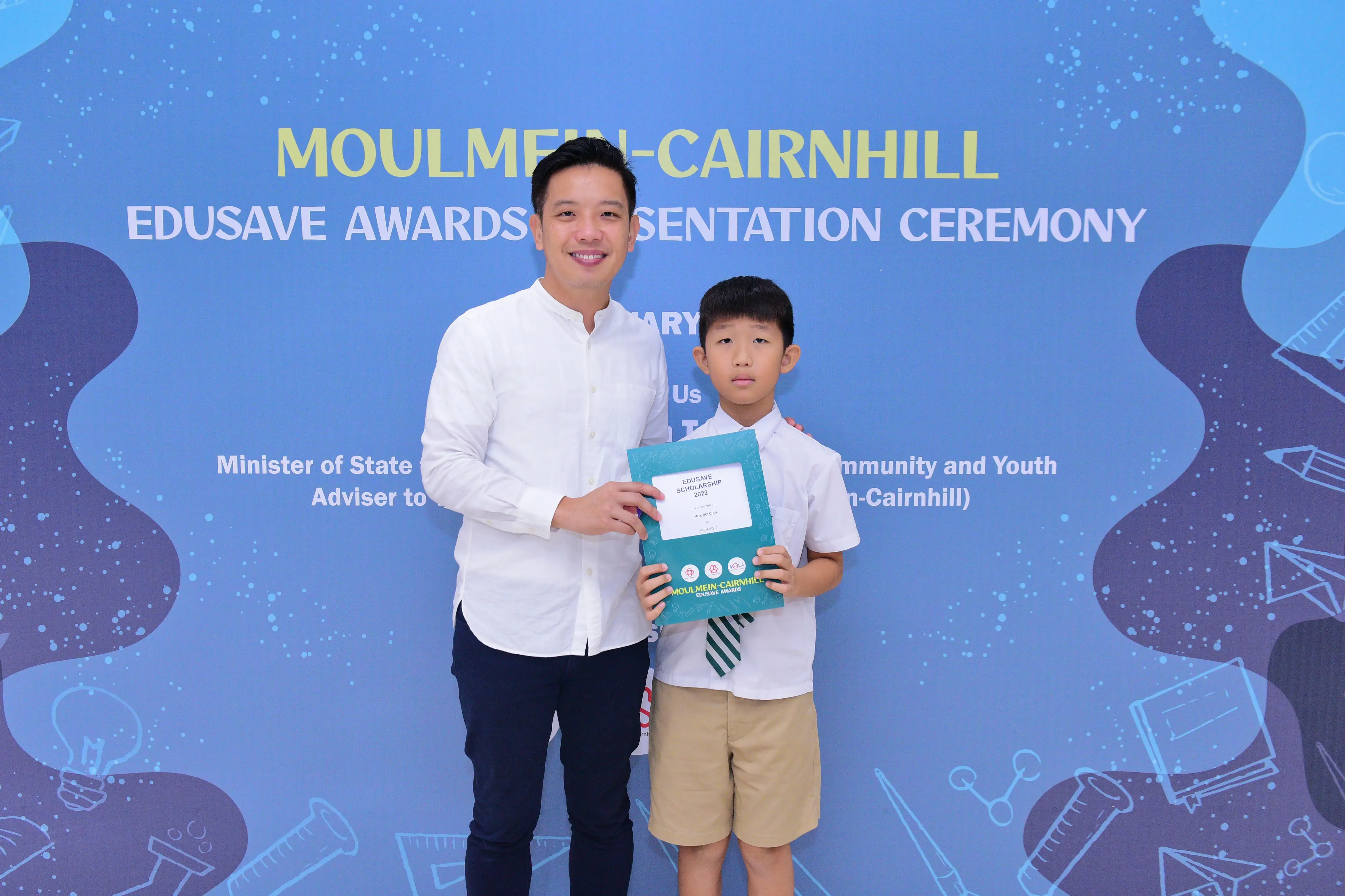 A man and a young boy hold a teal certificate folder at the Moulmein-Cairnhill Edusave Awards Presentation Ceremony.