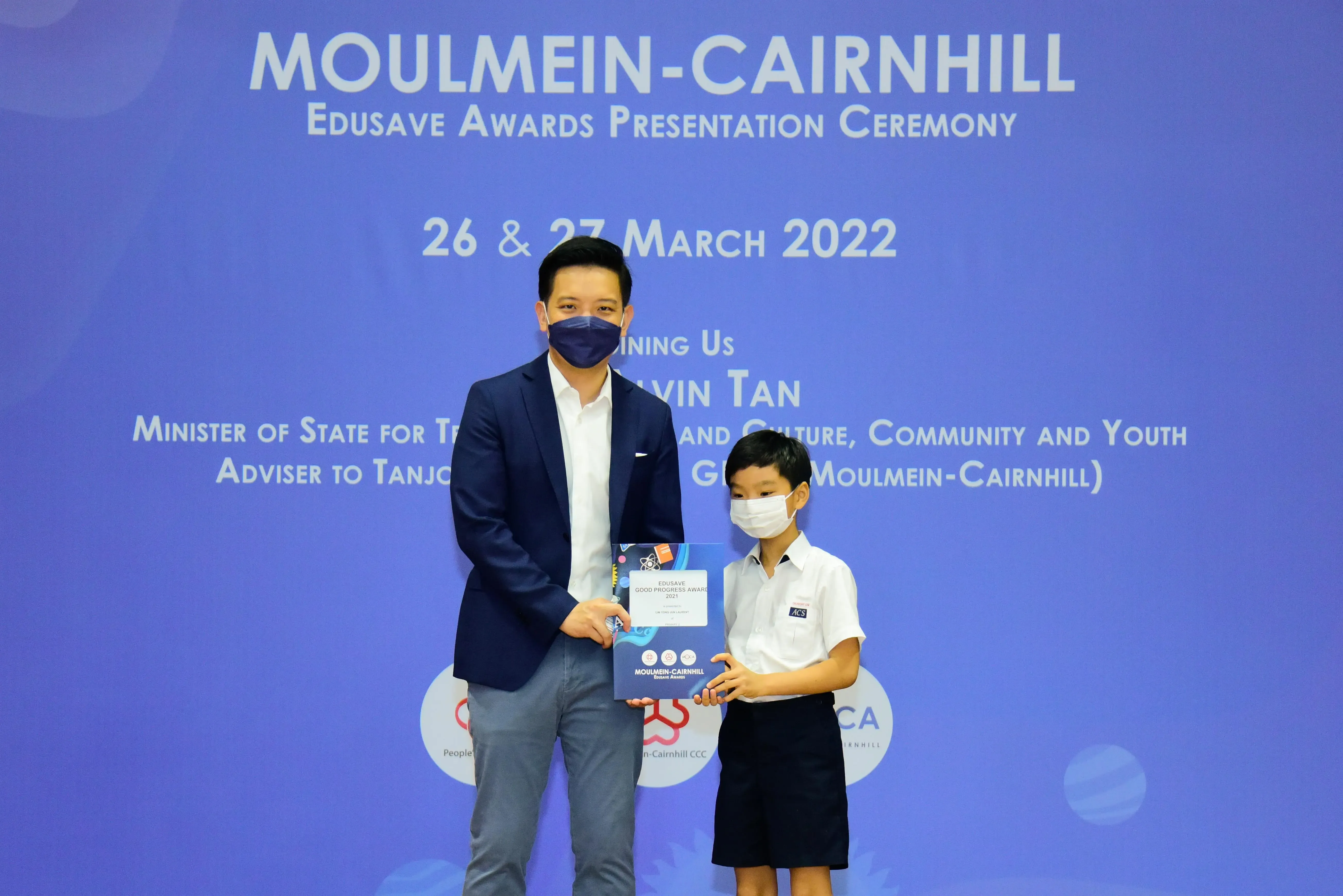 An adult and a young student in masks hold an award certificate at the Moulmein-Cairnhill Edusave Awards Ceremony 2022.