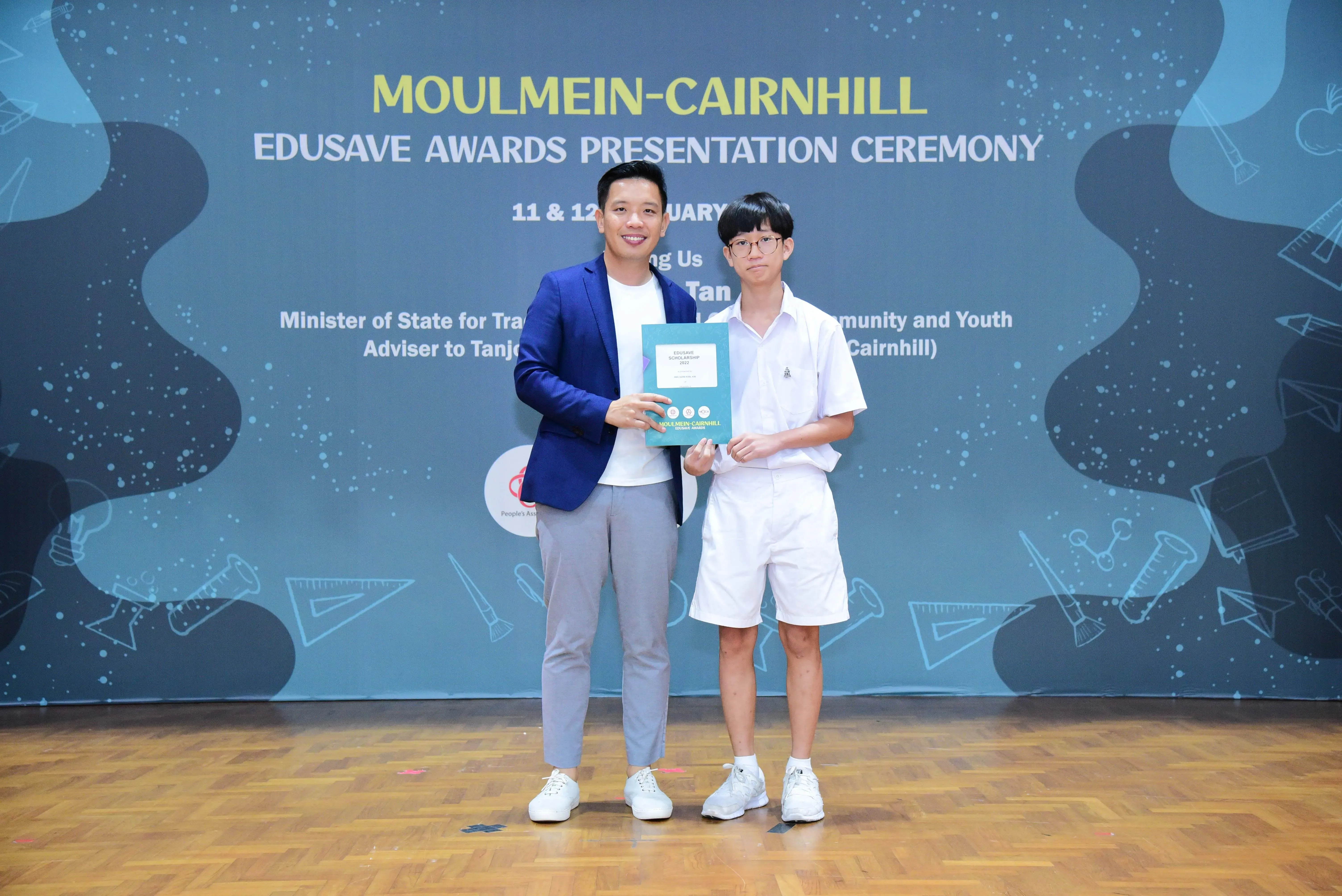 Two people pose holding a certificate at the Moulmein-Cairnhill Edusave Awards Presentation Ceremony backdrop.