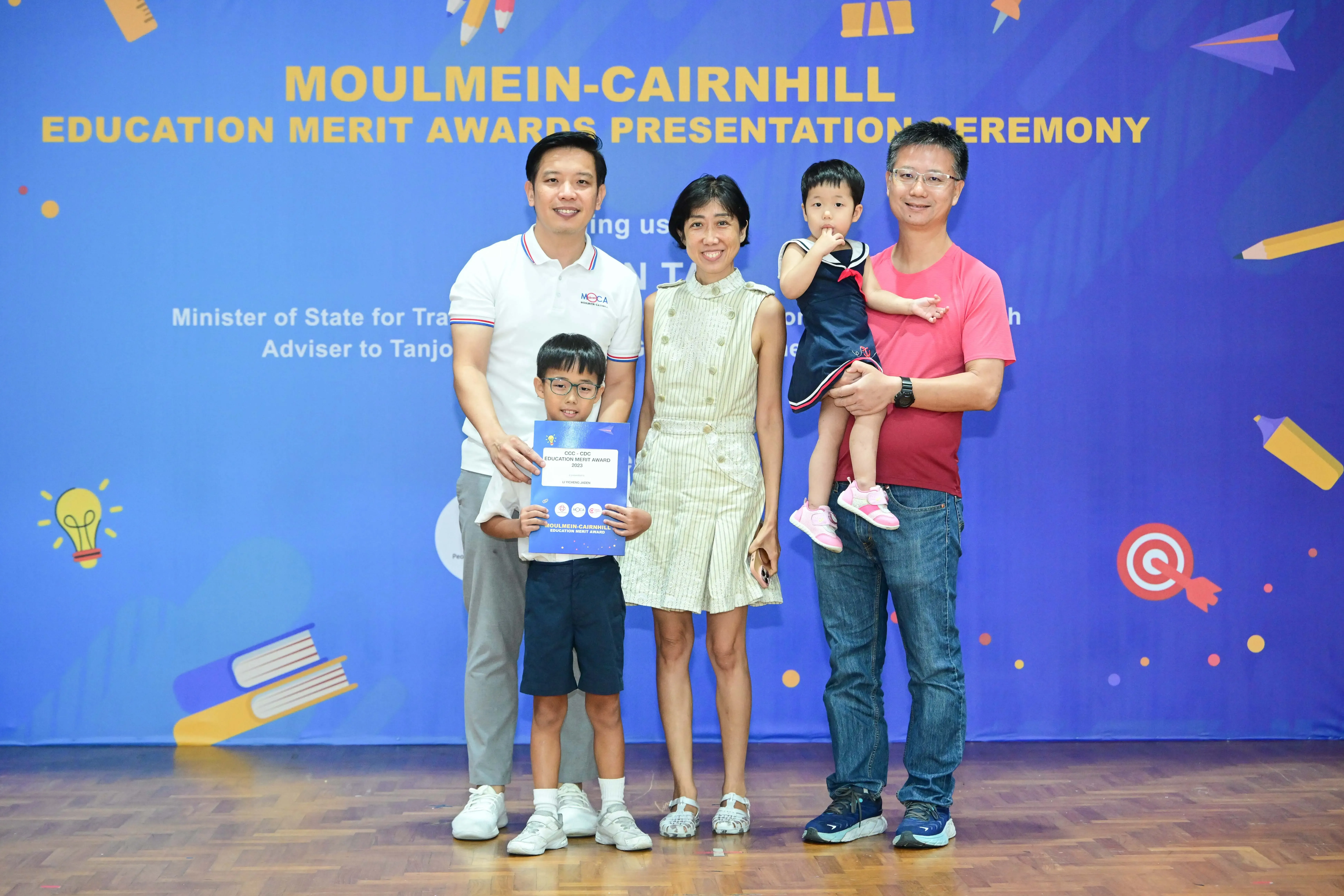 Four adults and two children pose at the Moulmein-Cairnhill Education Merit Awards Presentation Ceremony, boy holds certificate.