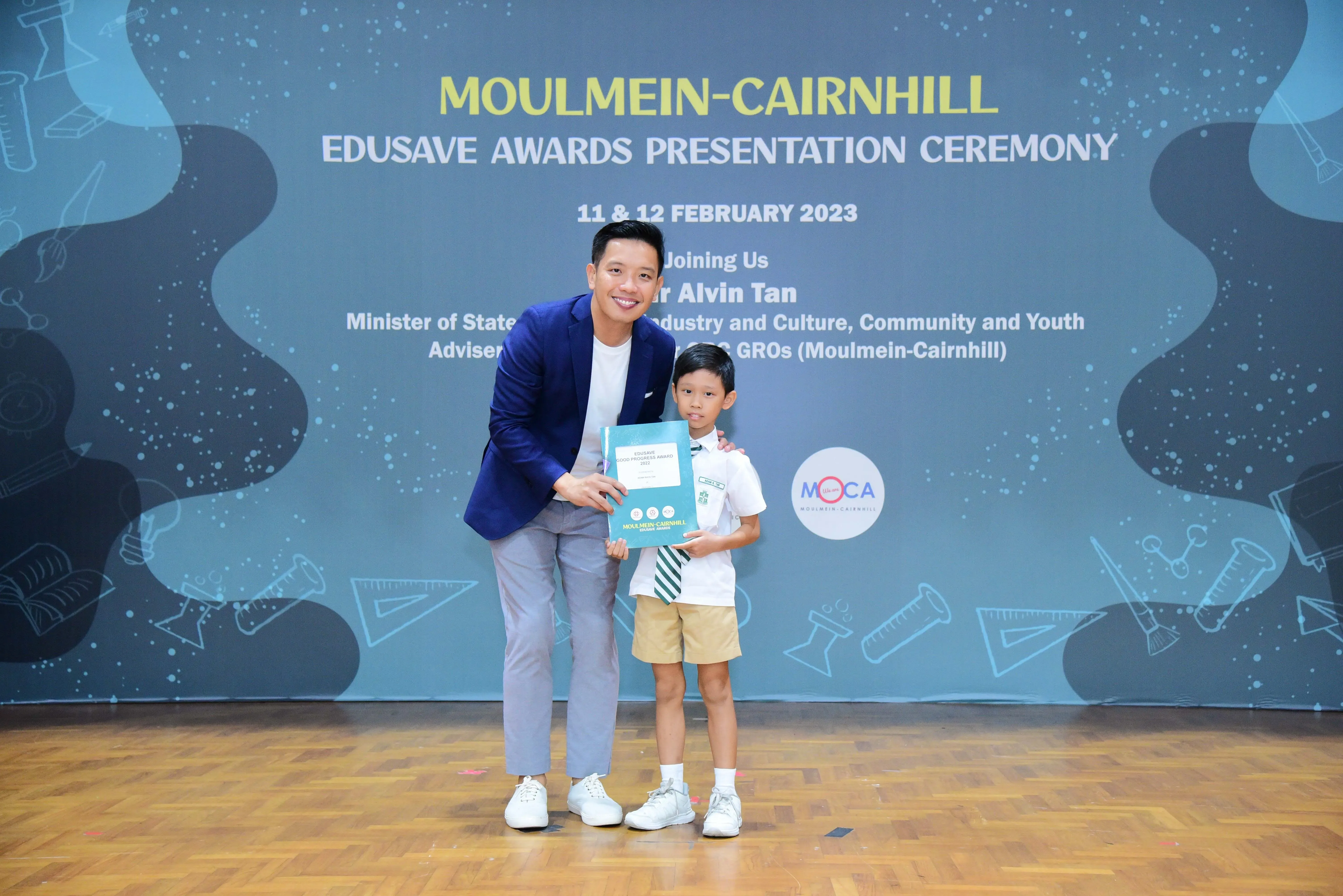 A man in a blue blazer and a young boy in school uniform hold a certificate at the Moulmein-Cairnhill Edusave Awards Ceremony.