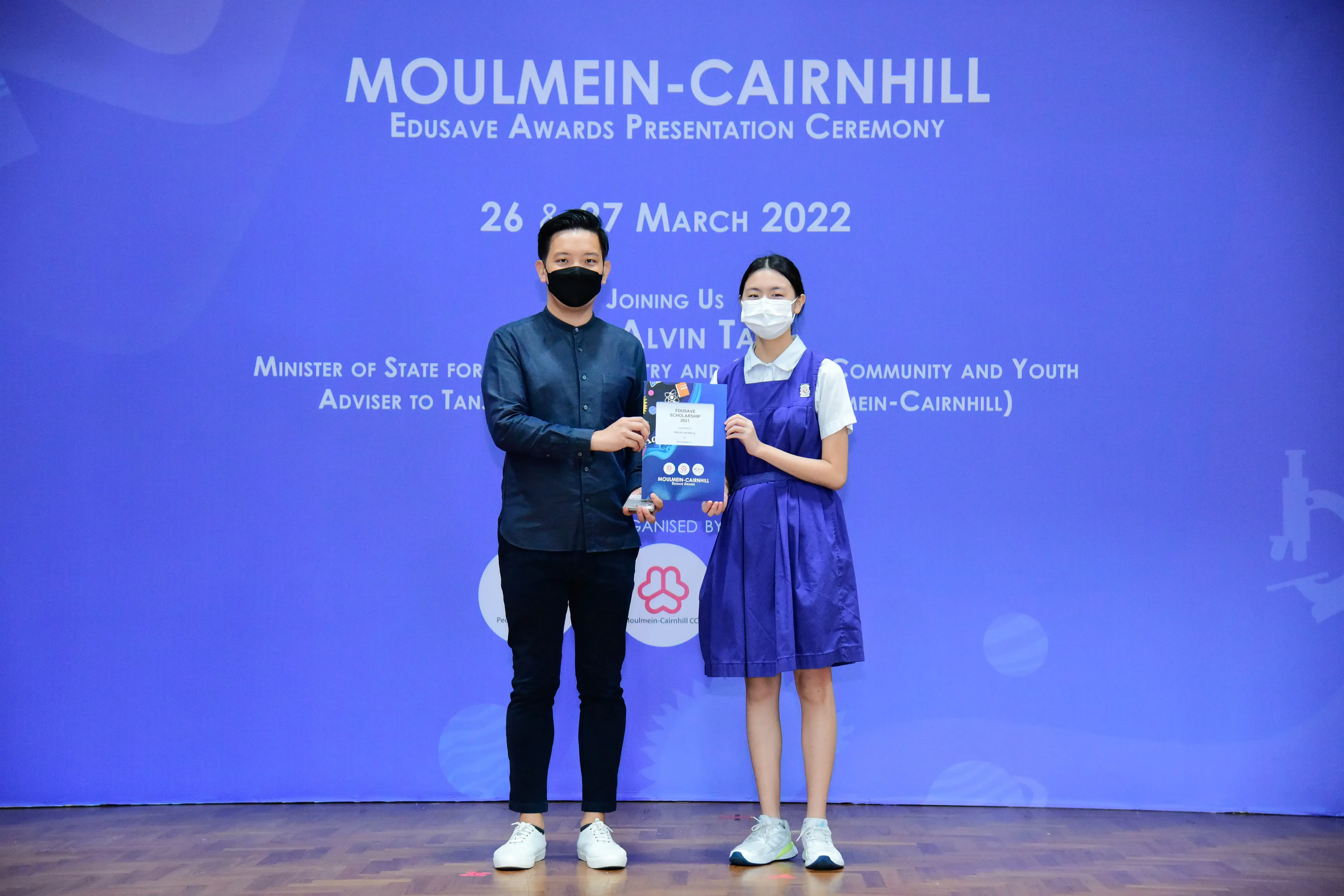 Two masked people hold an award booklet on stage at the Moulmein-Cairnhill Edusave Awards Ceremony, March 2022.