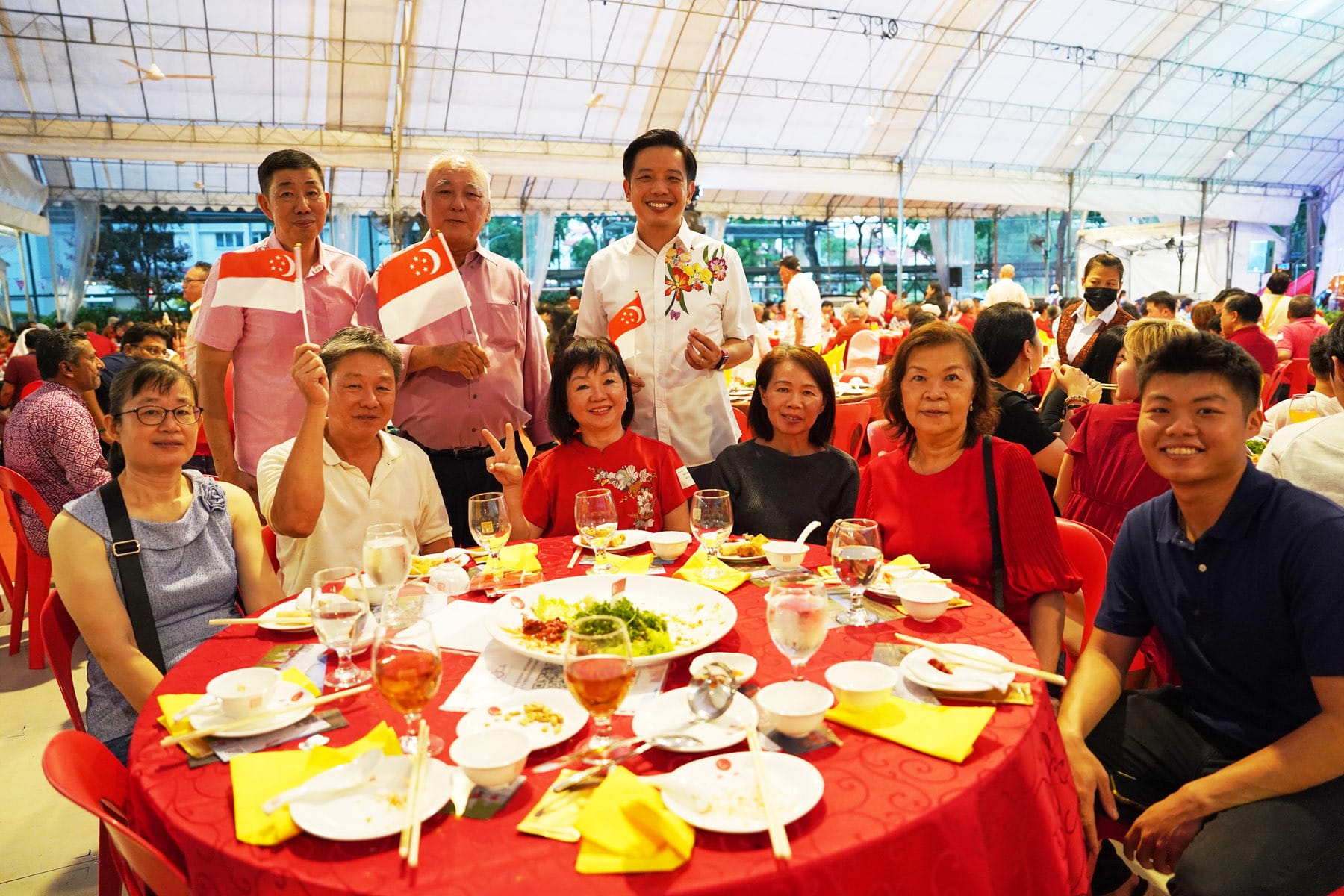 A group of smiling people pose at a red banquet table, some holding small Singapore flags at an outdoor celebration event.