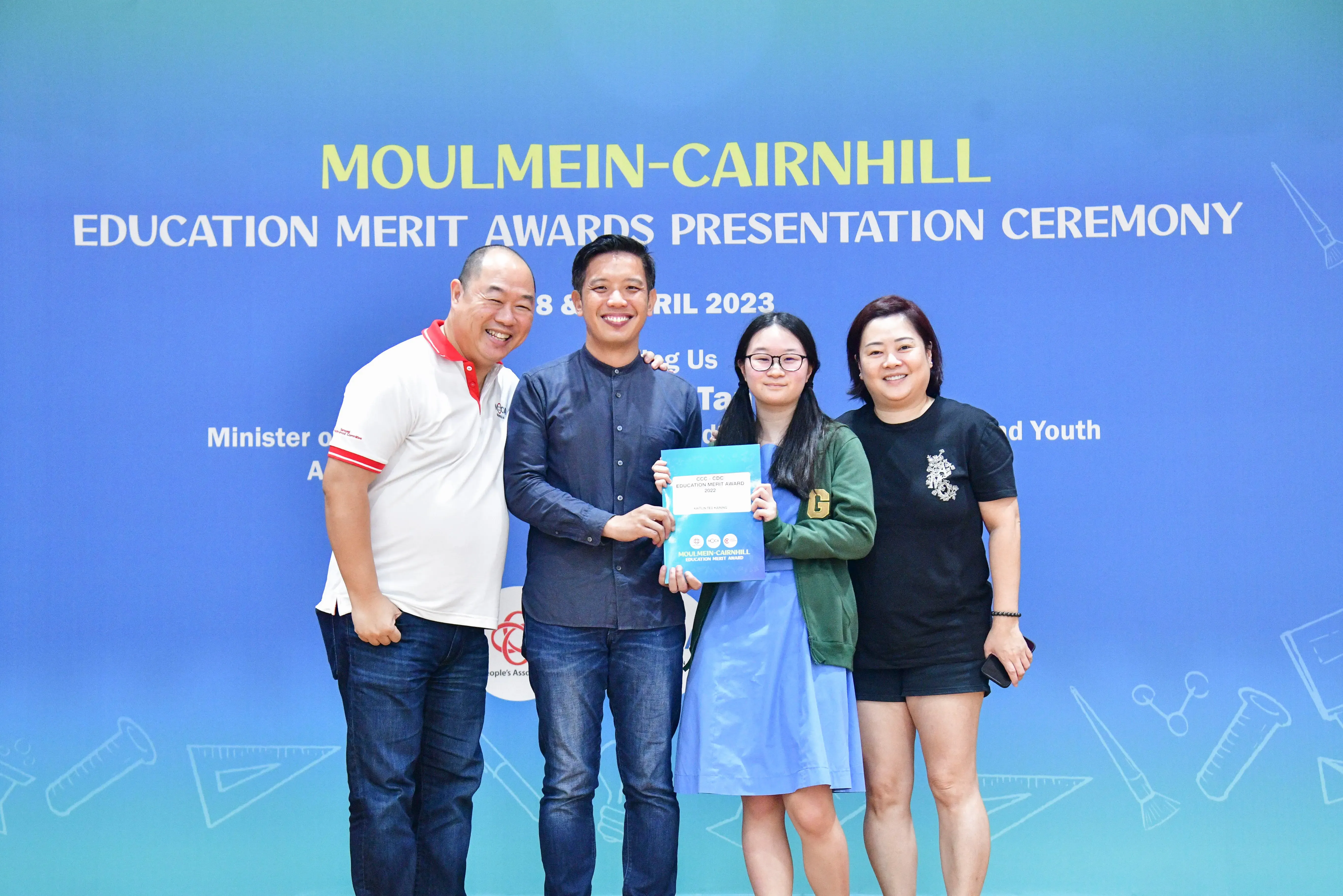 Four people pose smiling before a Moulmein-Cairnhill Education Merit Awards 2023 banner, one holding a certificate.