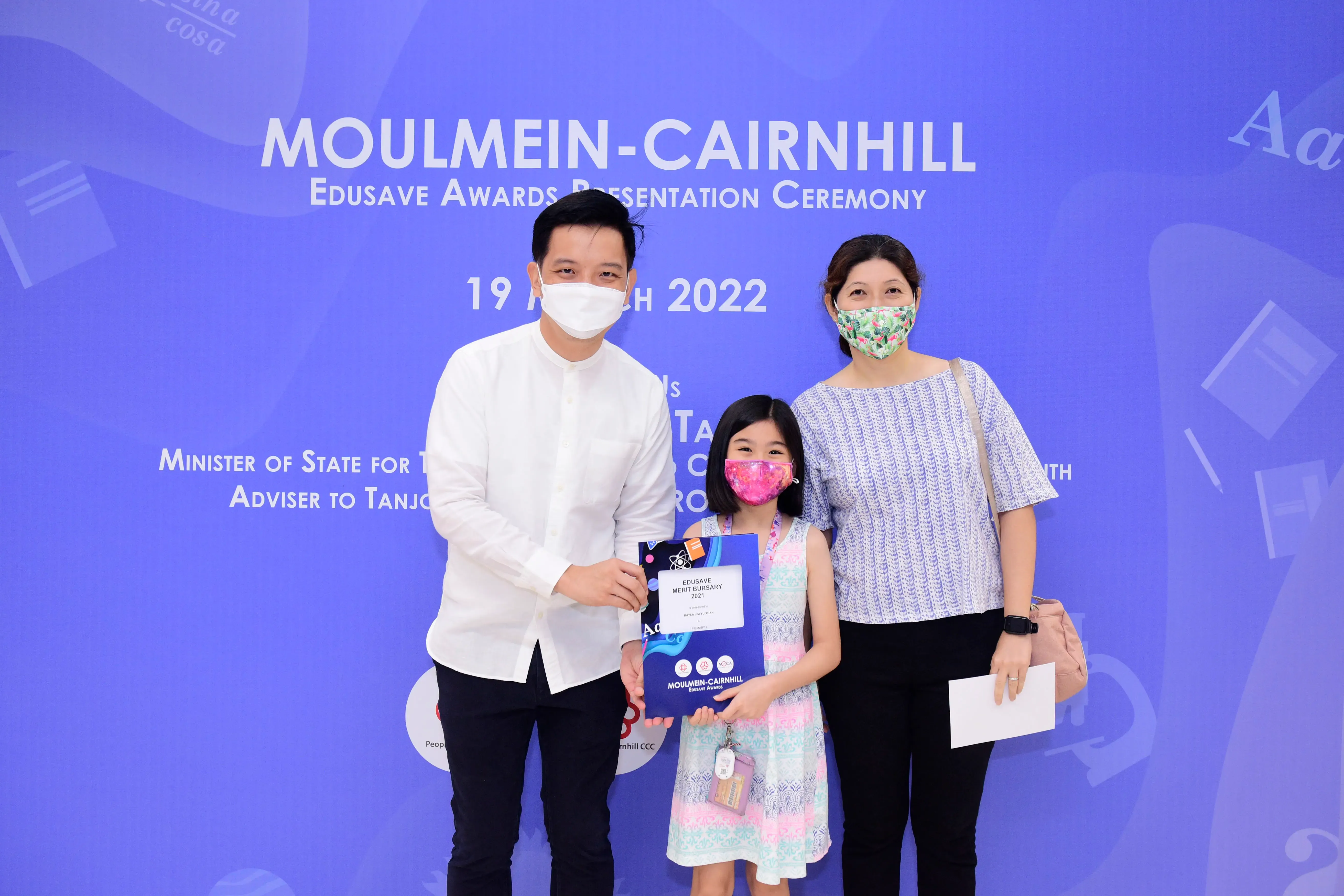 Two masked adults and a masked girl holding an Edusave Merit Bursary certificate at a 2022 awards ceremony backdrop.