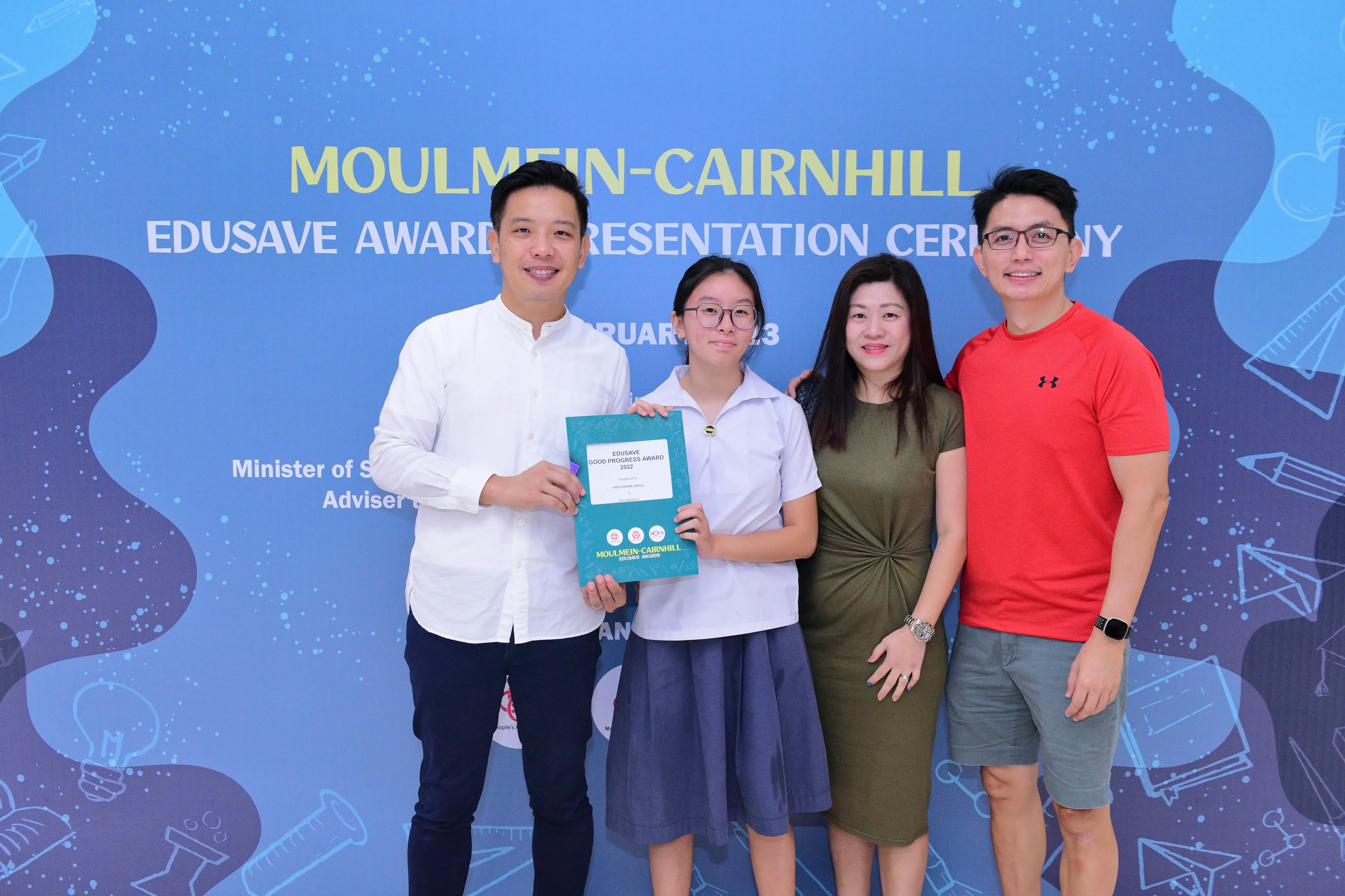 Four people pose smiling before a Moulmein-Cairnhill Edusave Award Presentation Ceremony banner, girl holding a certificate.
