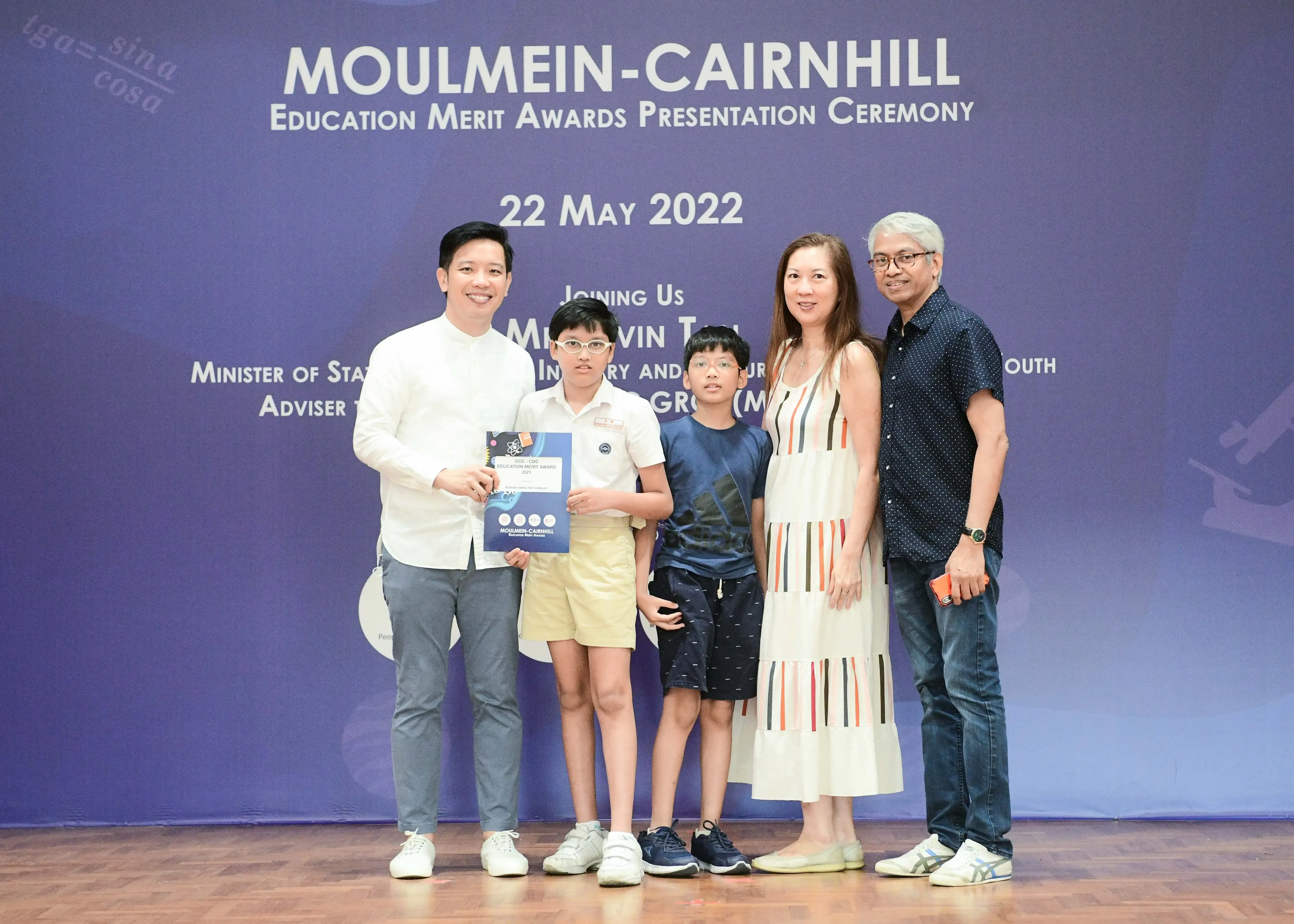 Five people pose before a Moulmein-Cairnhill Education Merit Awards banner dated 22 May 2022.