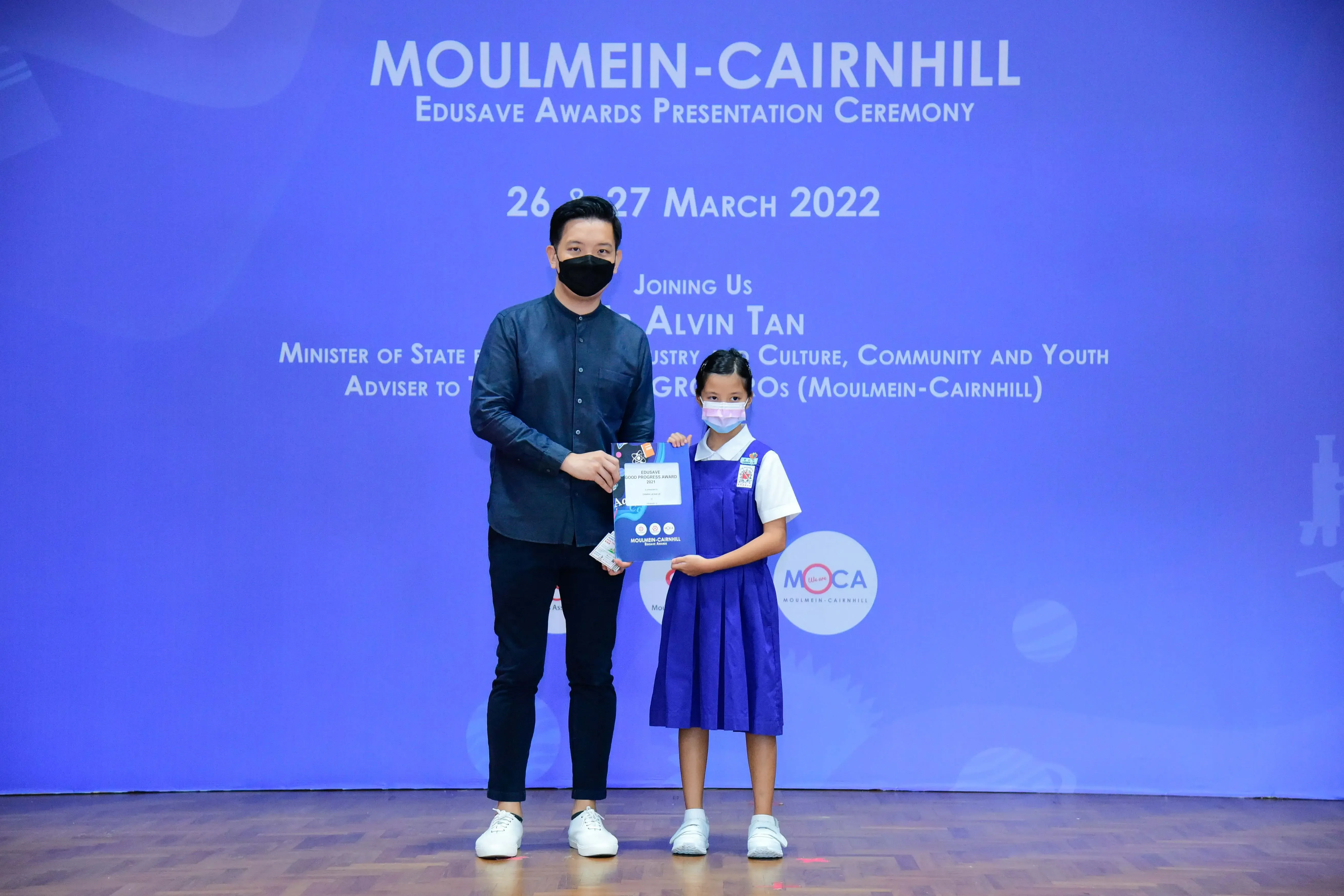 A masked man and a young girl in school uniform hold an award together at the Moulmein-Cairnhill Edusave ceremony.