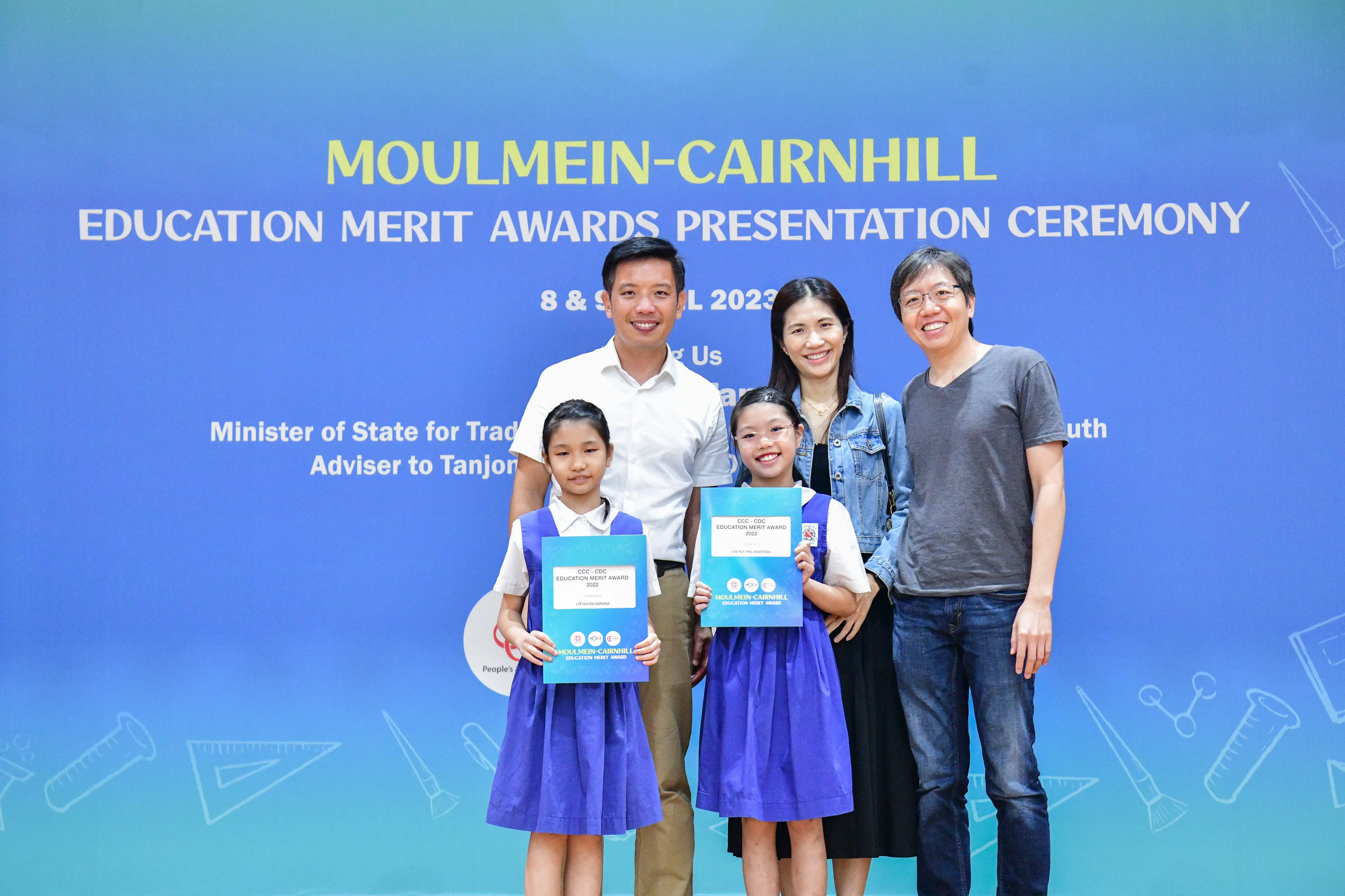 Two girls in blue uniforms hold merit award certificates, posing with three adults at the Moulmein-Cairnhill Education Merit Awards 2023.