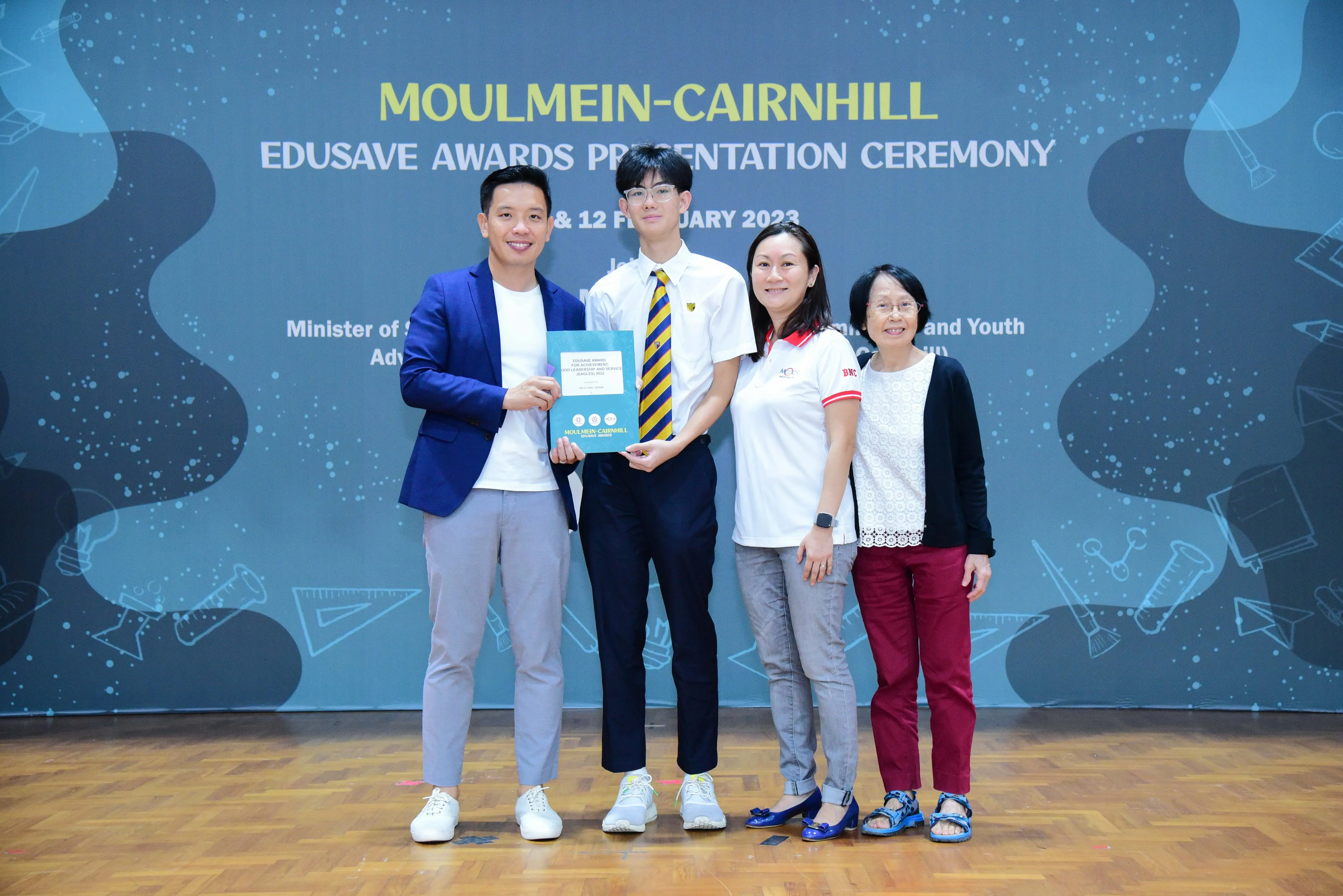Four people pose at the Moulmein-Cairnhill Edusave Awards Presentation Ceremony, with a student holding a certificate.