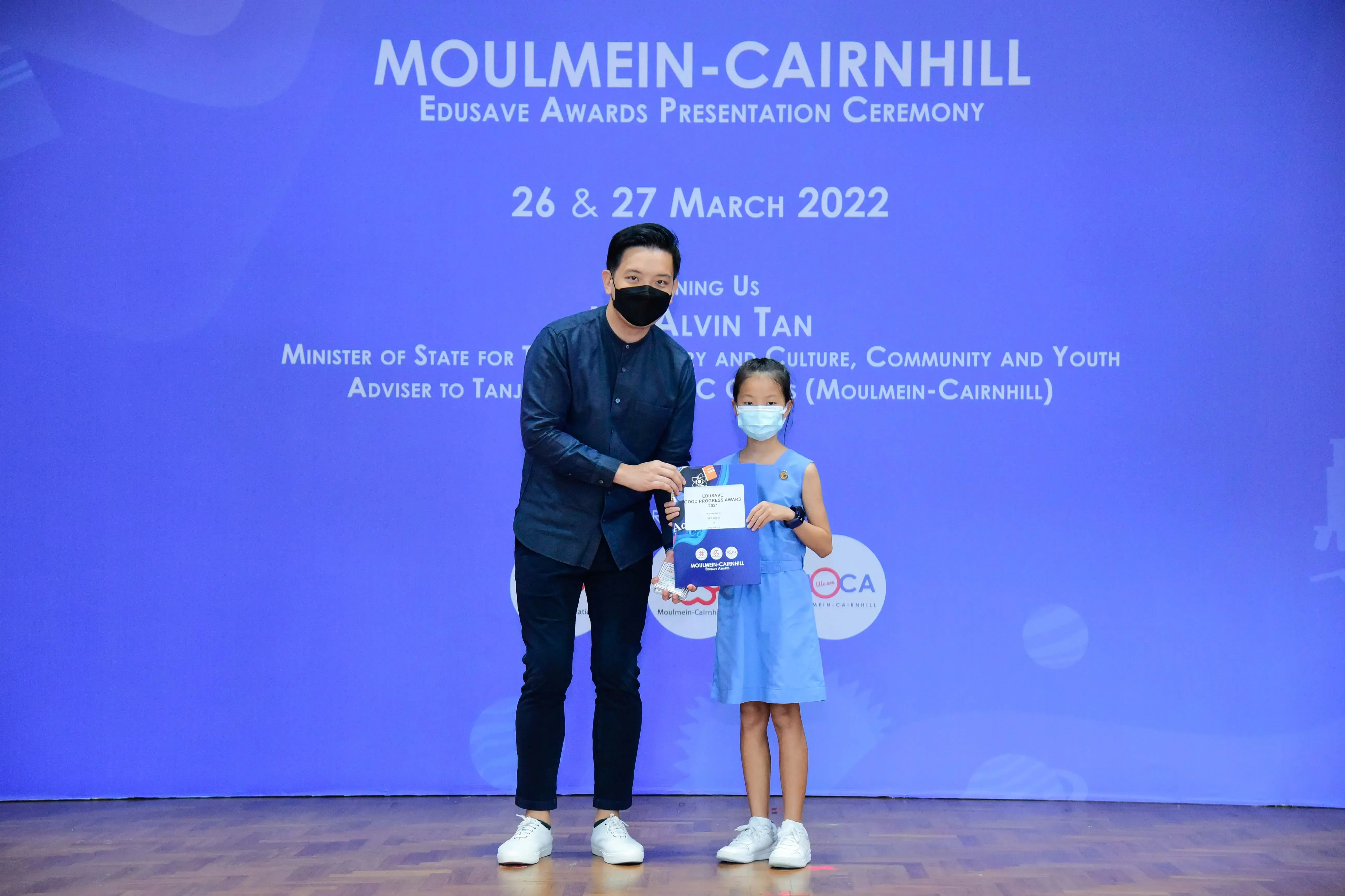 An adult and a young girl wearing masks hold an award certificate at the Moulmein-Cairnhill Edusave Awards Ceremony 2022.