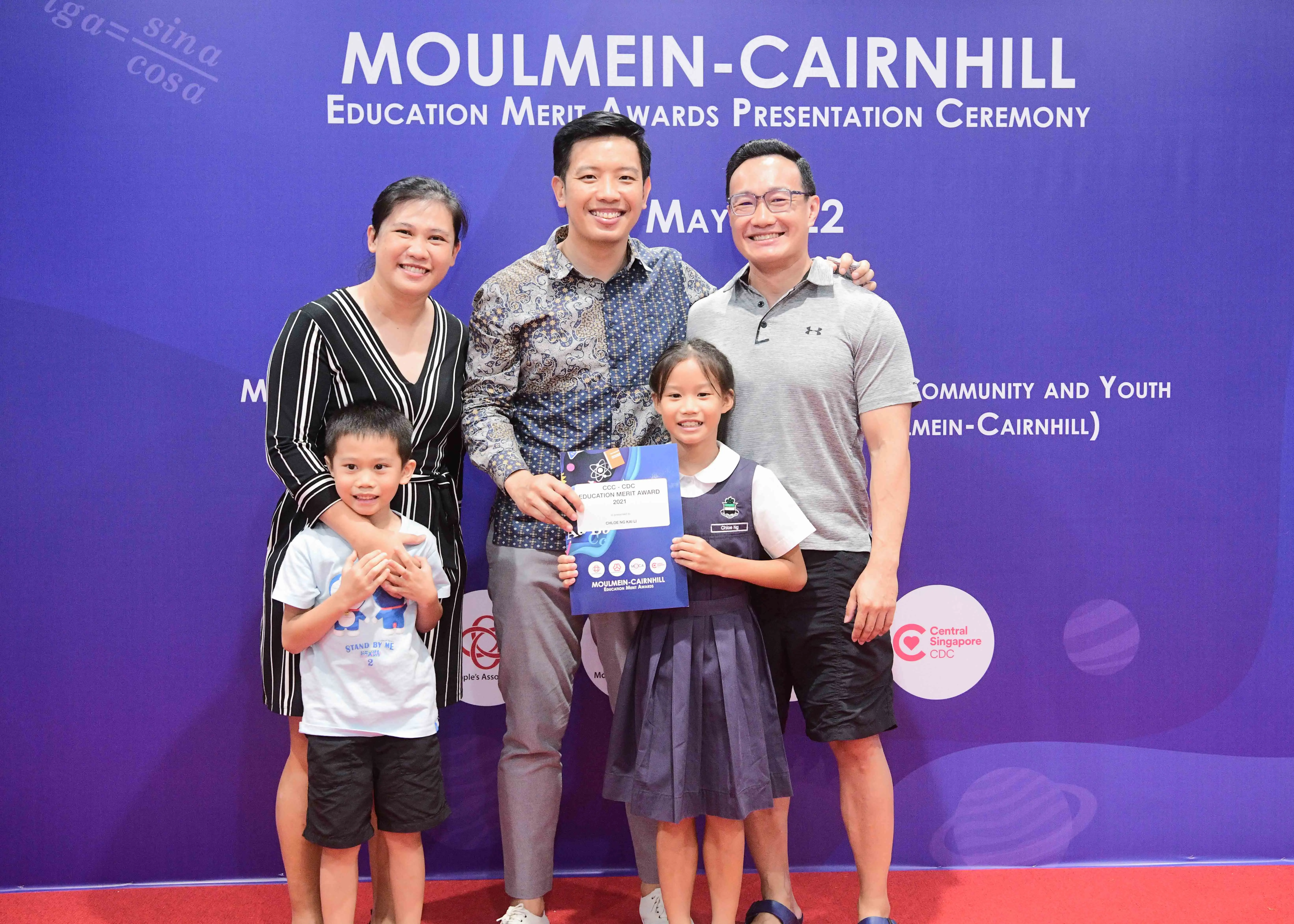 Five people pose smiling before a Moulmein-Cairnhill Education Merit Awards Presentation Ceremony banner, girl holds certificate.