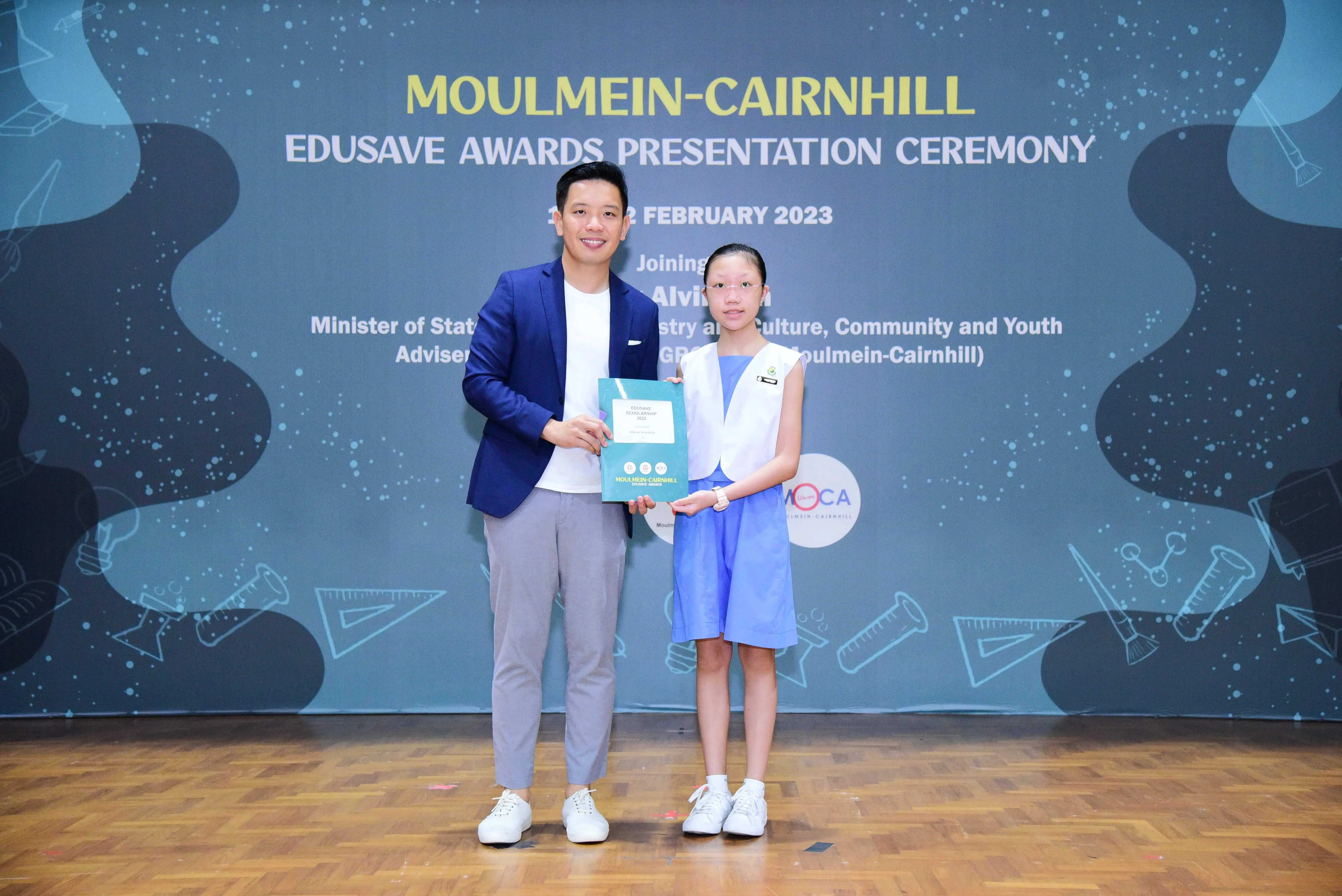 A man and a young girl hold a certificate on stage at the Moulmein-Cairnhill Edusave Awards Ceremony, February 2023.