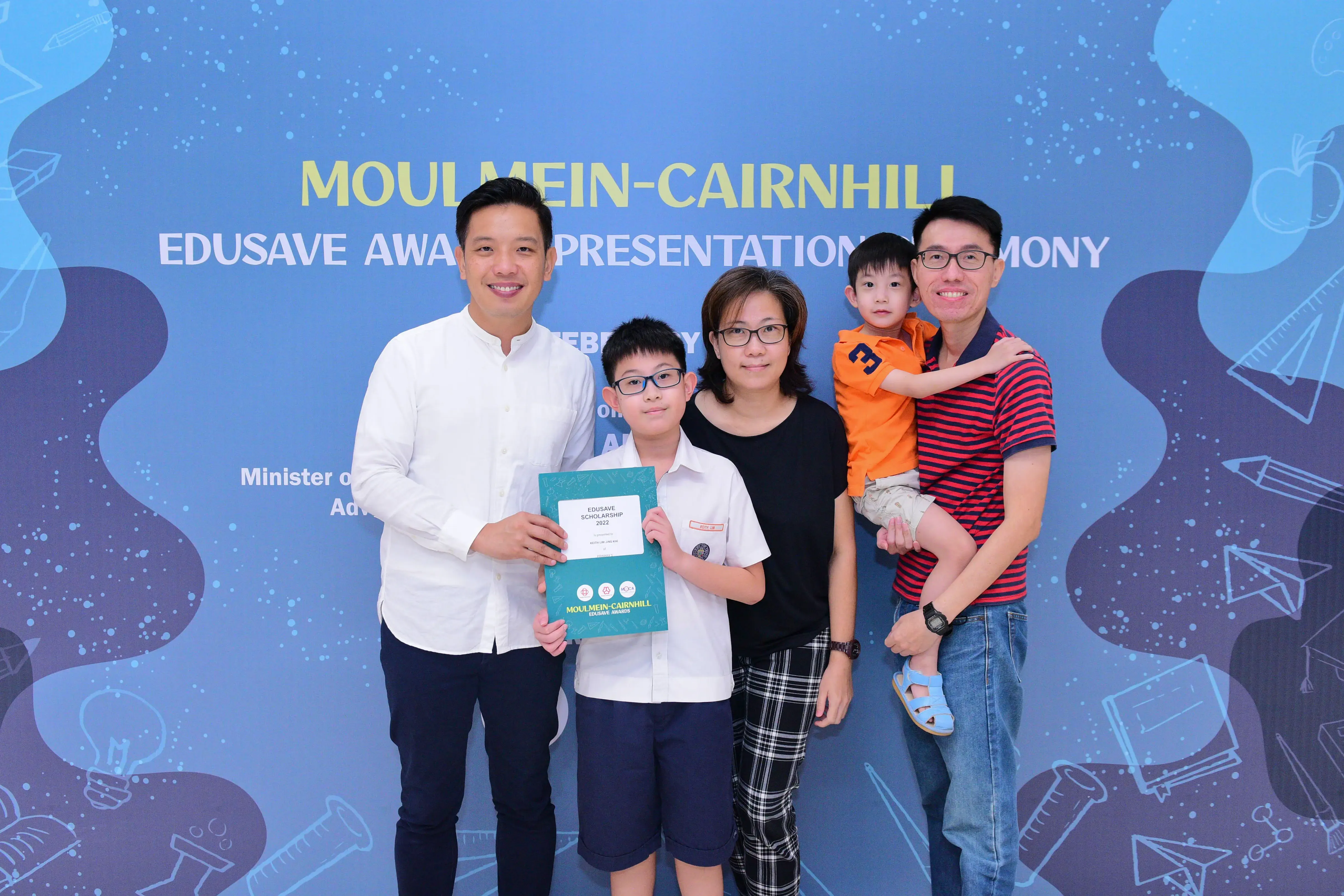 Family of four poses with a boy holding a 2022 Edusave Scholarship certificate at Moulmein-Cairnhill award ceremony.