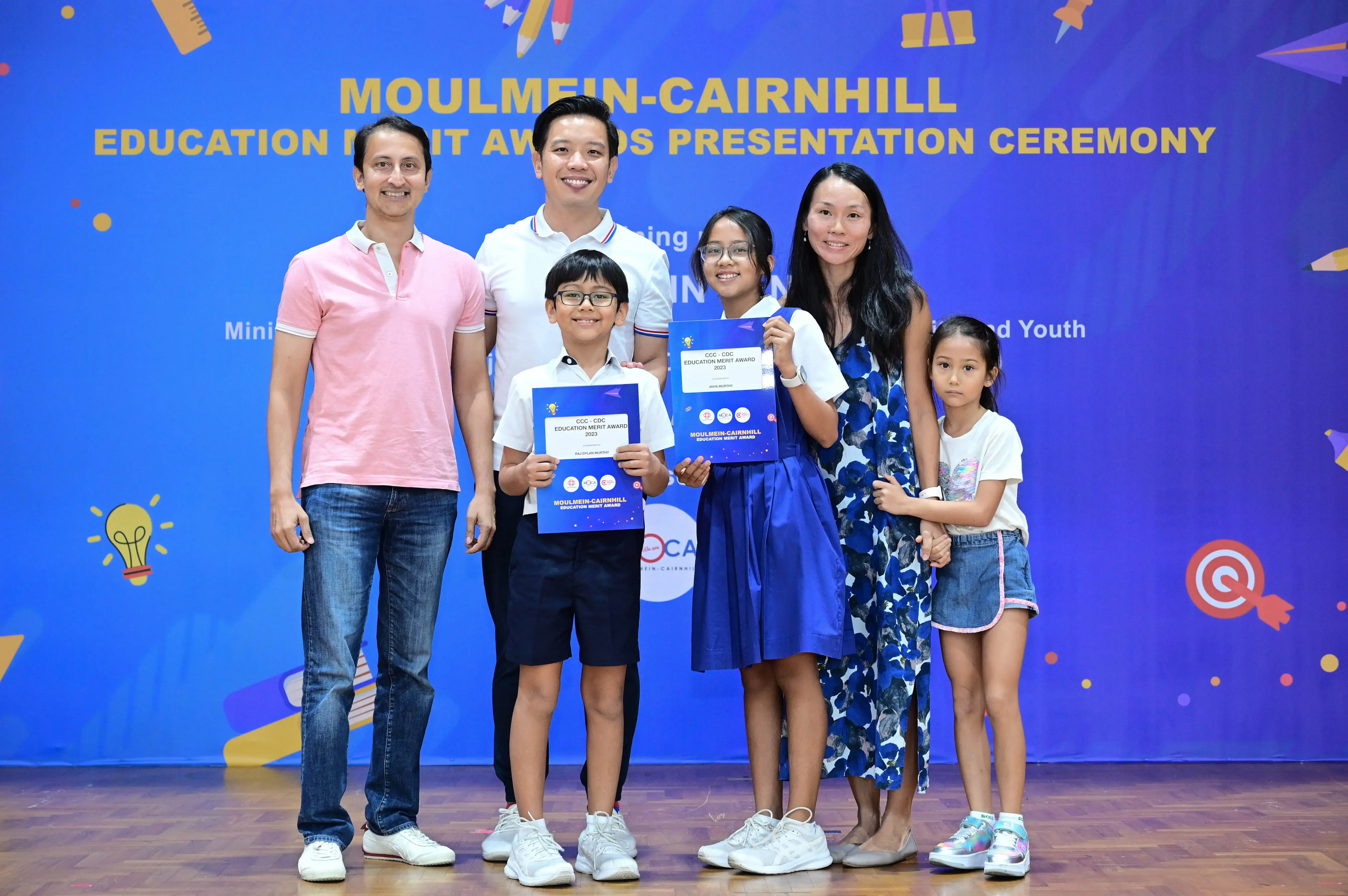Six people pose at the Moulmein-Cairnhill Education Merit Awards ceremony, two children holding blue award certificates.