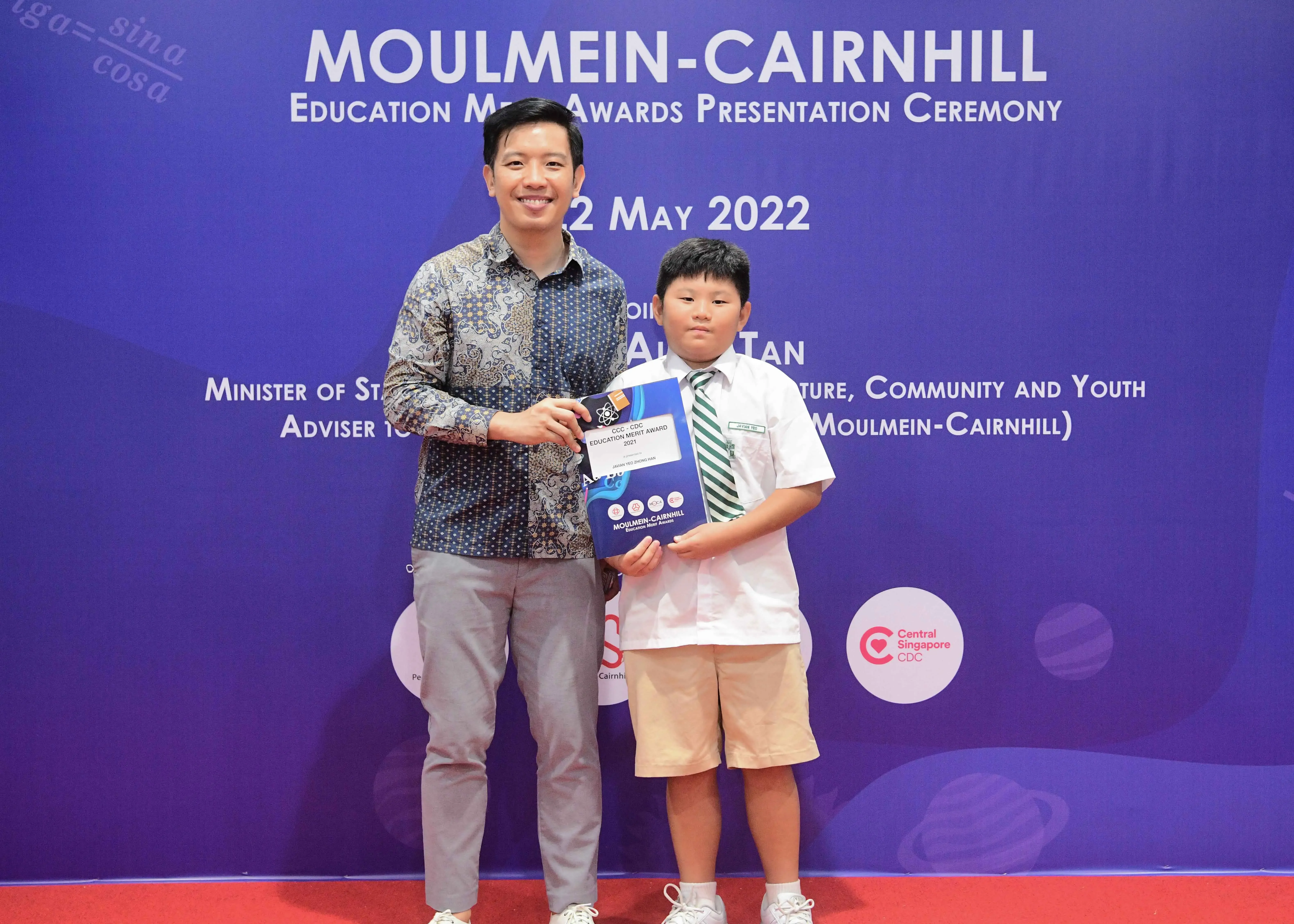 An adult and a young boy hold an award certificate at the Moulmein-Cairnhill Education Merit Awards ceremony, May 2022.
