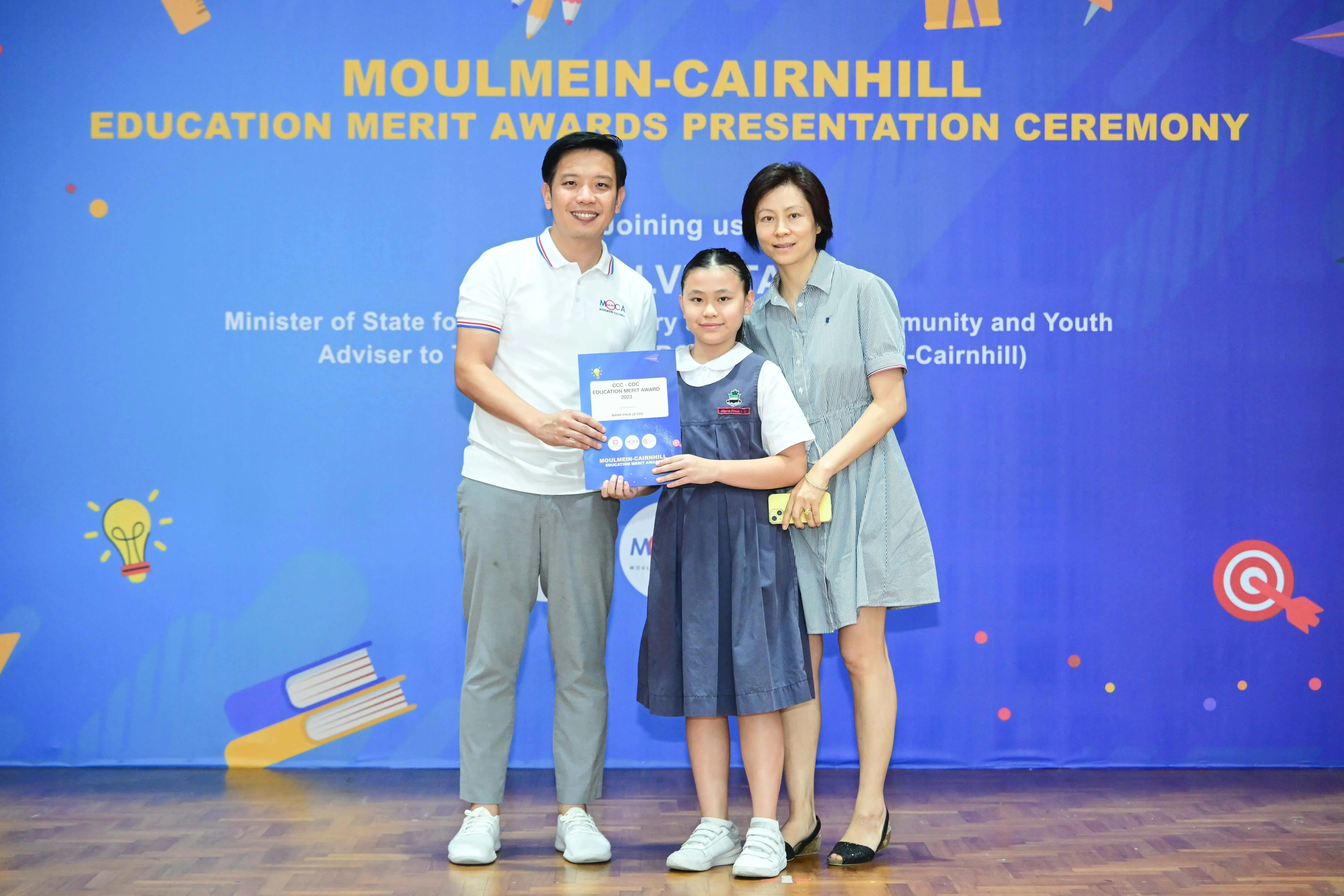 A girl in school uniform holds an award certificate flanked by two adults at the Moulmein-Cairnhill Education Merit Awards.
