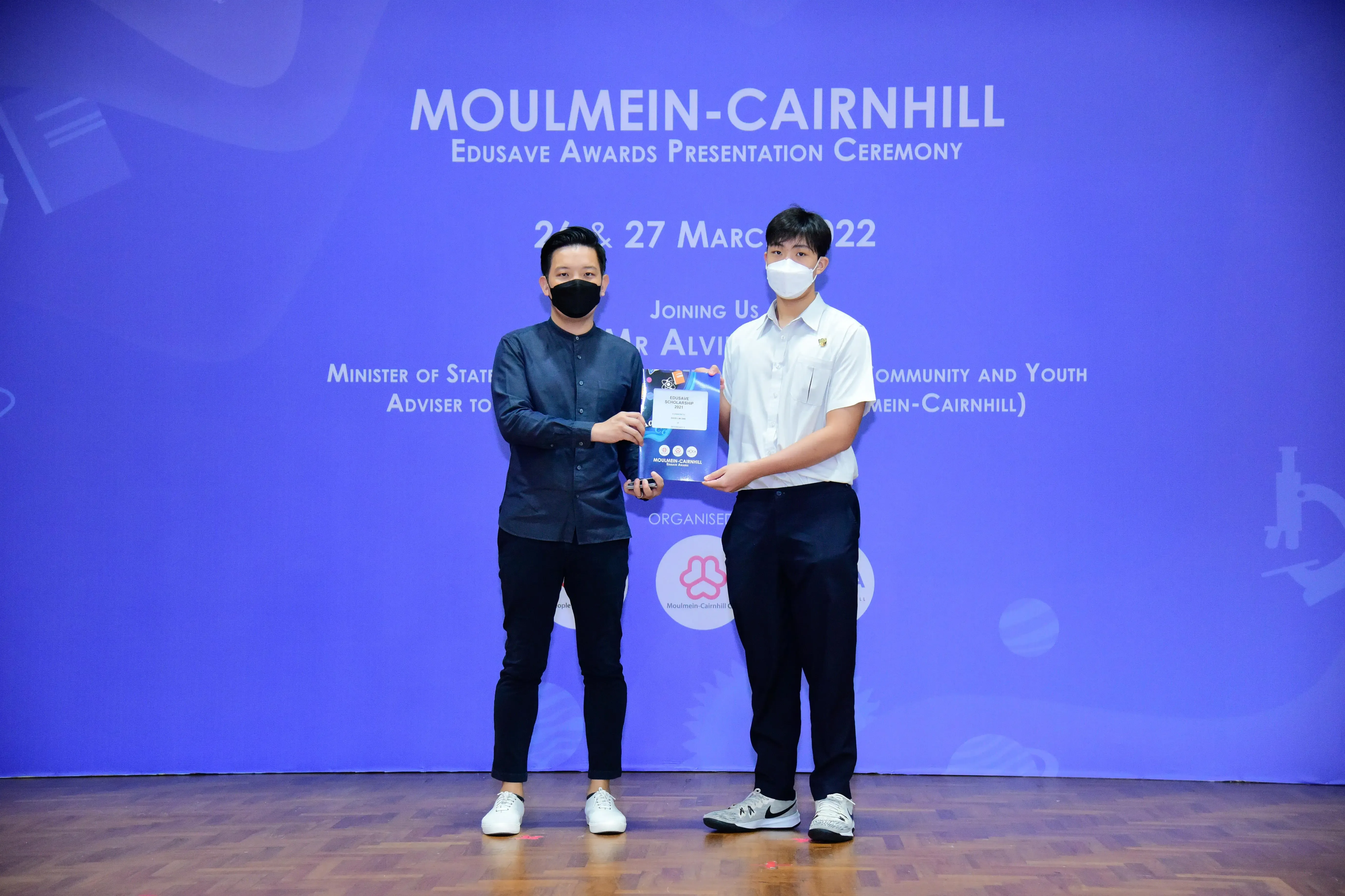 Two masked men hold a certificate at the Moulmein-Cairnhill Edusave Awards Presentation Ceremony, March 2022.