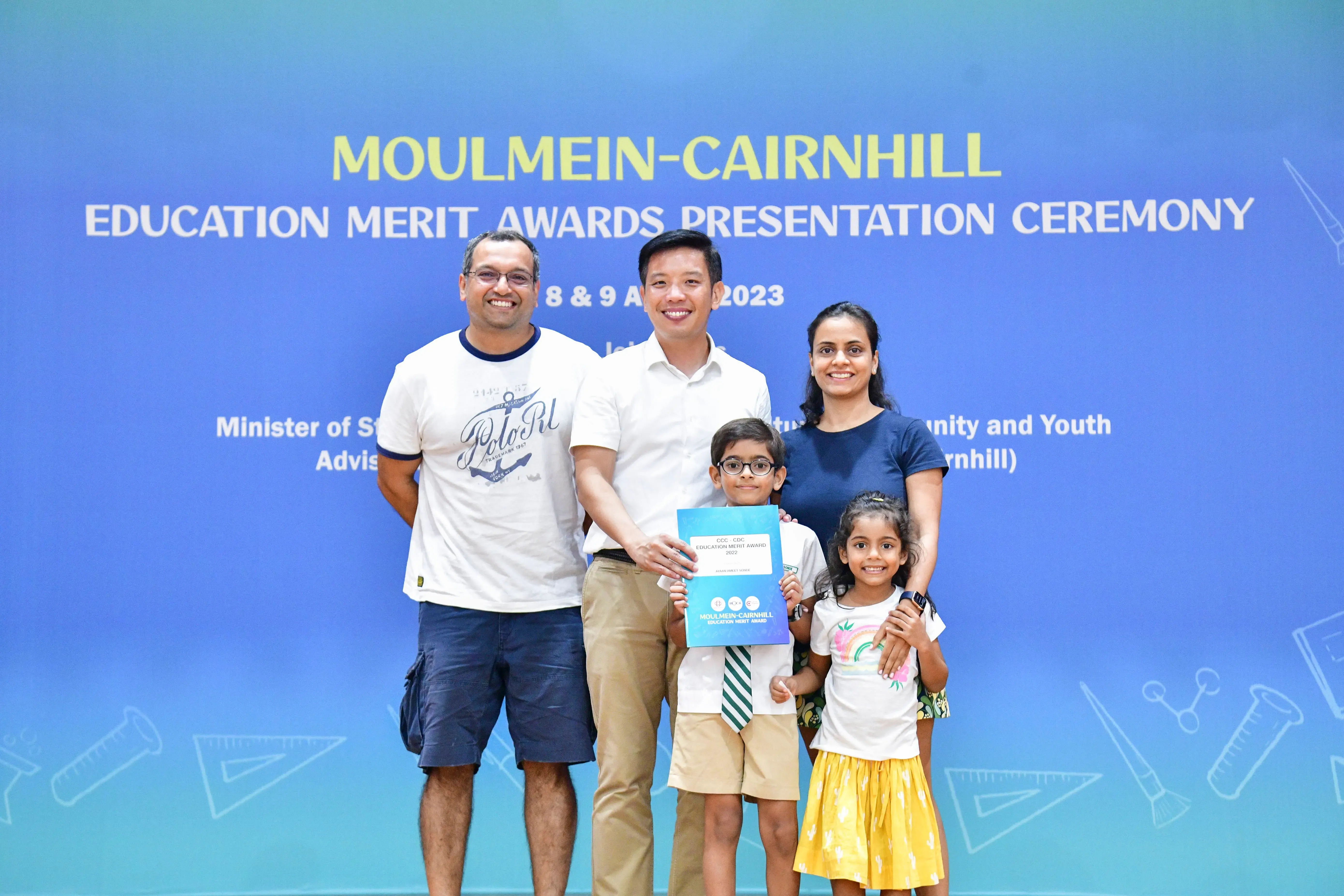 Five people pose before a Moulmein-Cairnhill Education Merit Awards 2023 banner, with a boy holding a certificate.