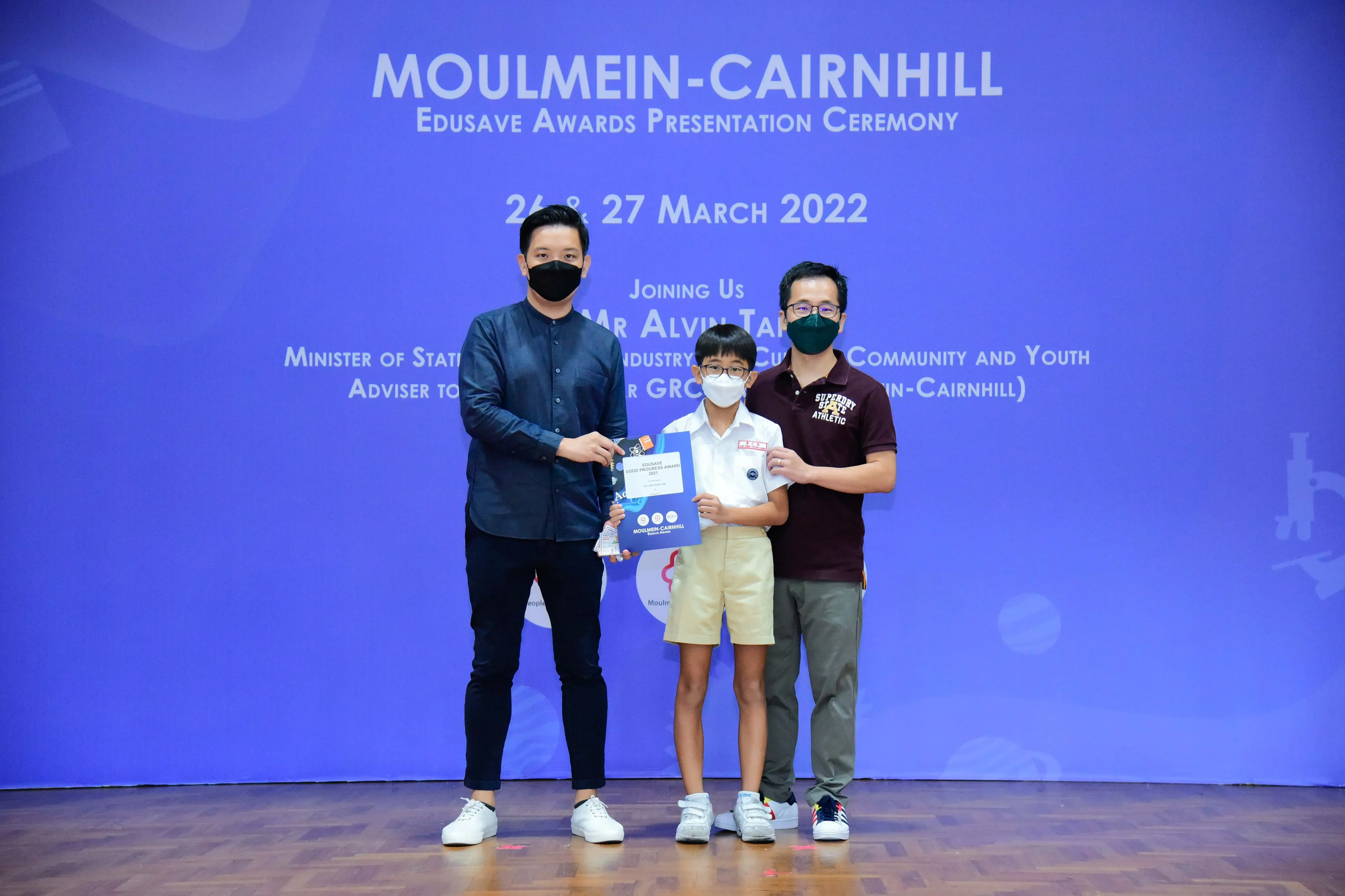 Three masked people pose at the Moulmein-Cairnhill Edusave Awards Ceremony on 26-27 March 2022, with a child holding an award.