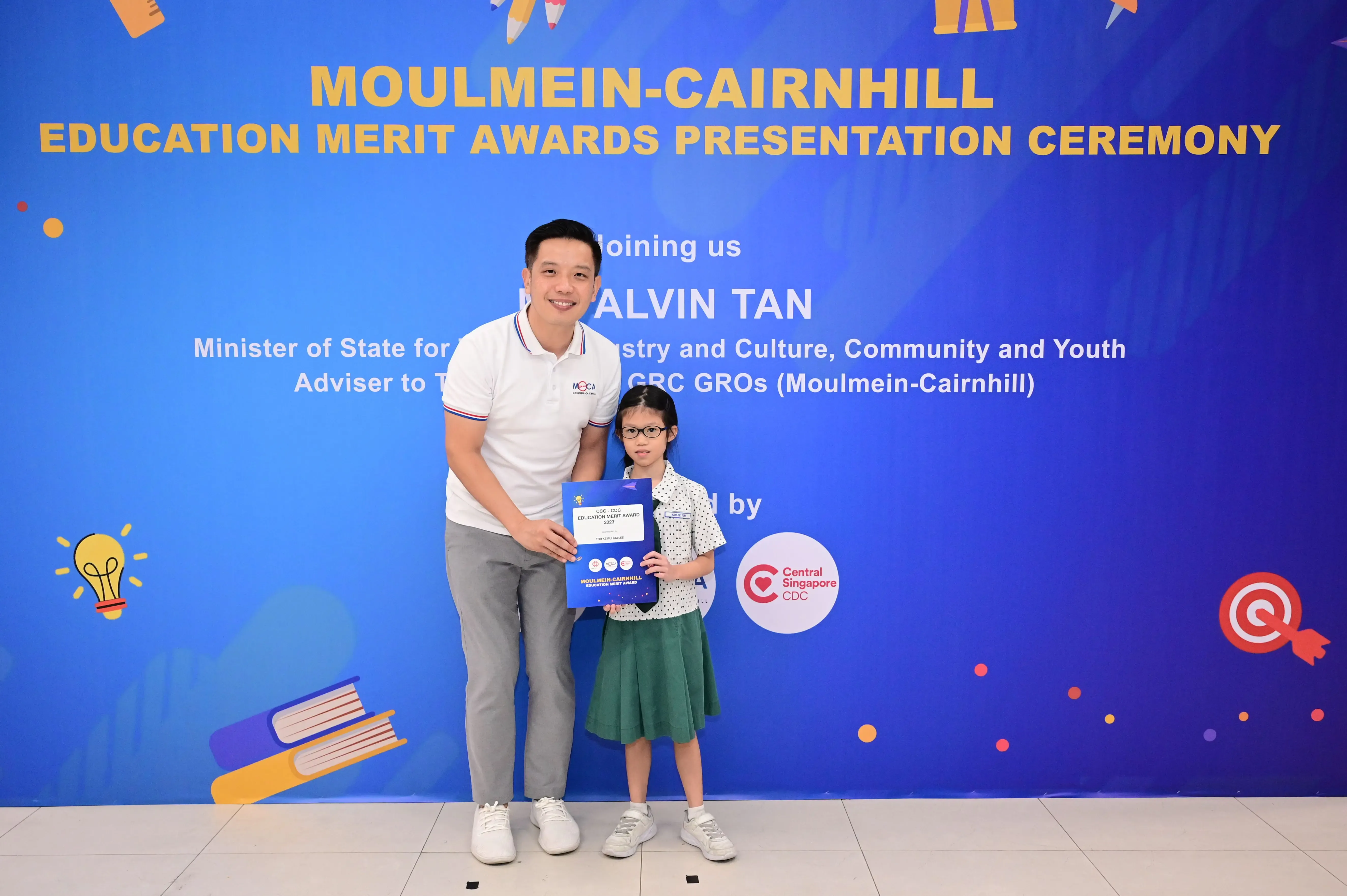 A man and a young girl holding an award certificate stand before a Moulmein-Cairnhill Education Merit Awards banner.