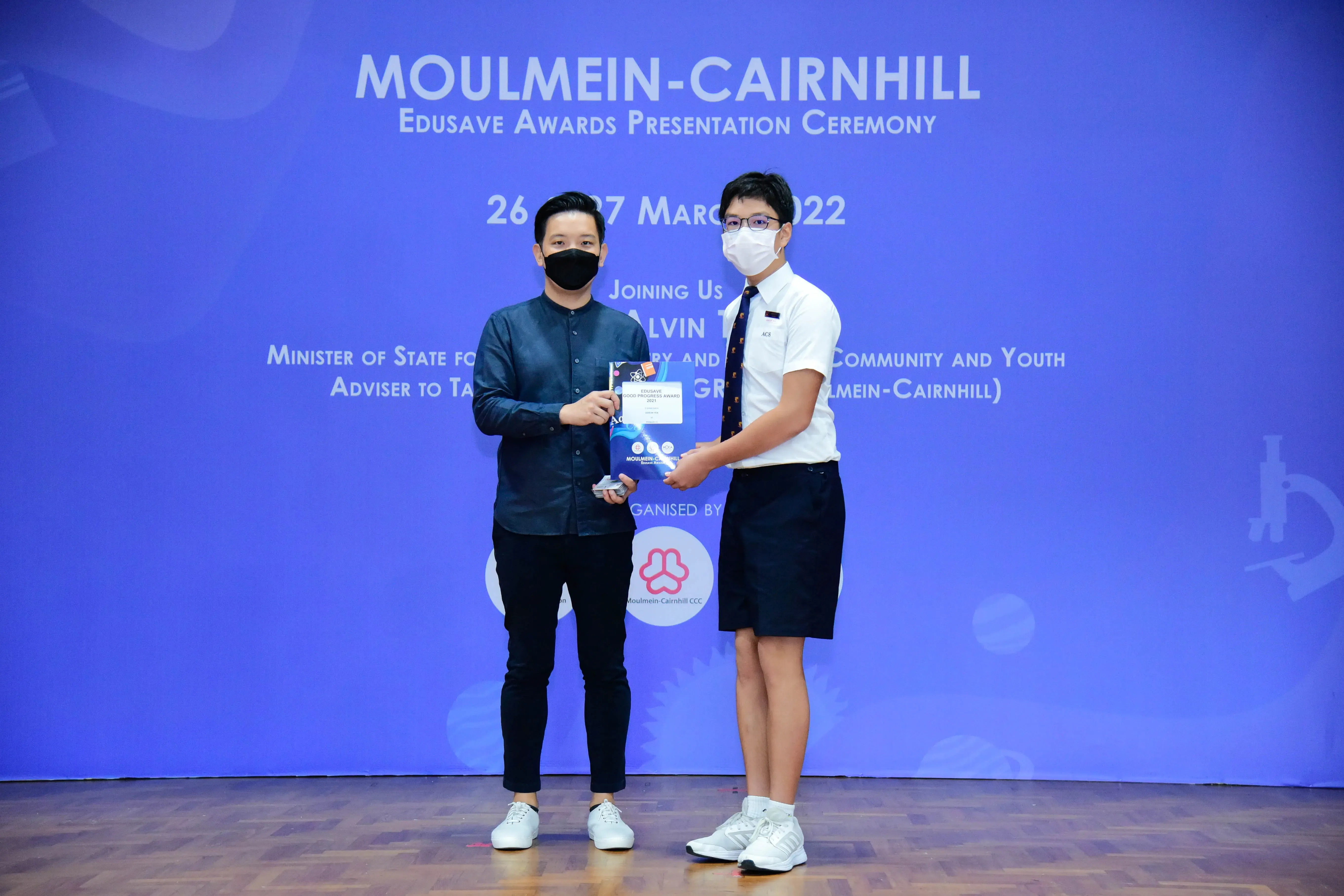 Two masked men hold an award certificate on stage at the Moulmein-Cairnhill Edusave Awards Ceremony, March 2022.