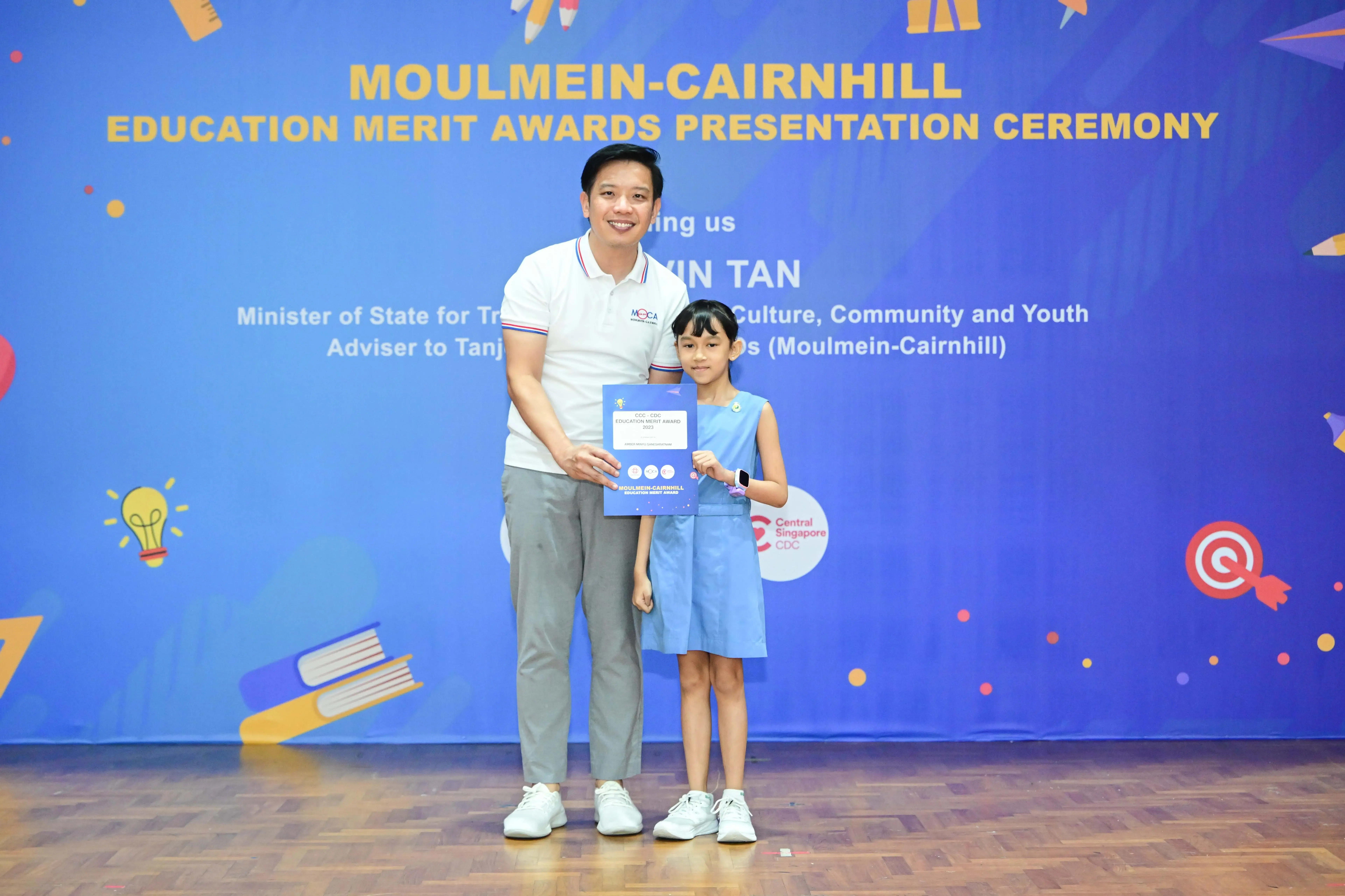A man and a young girl hold a certificate in front of a Moulmein-Cairnhill Education Merit Awards banner.