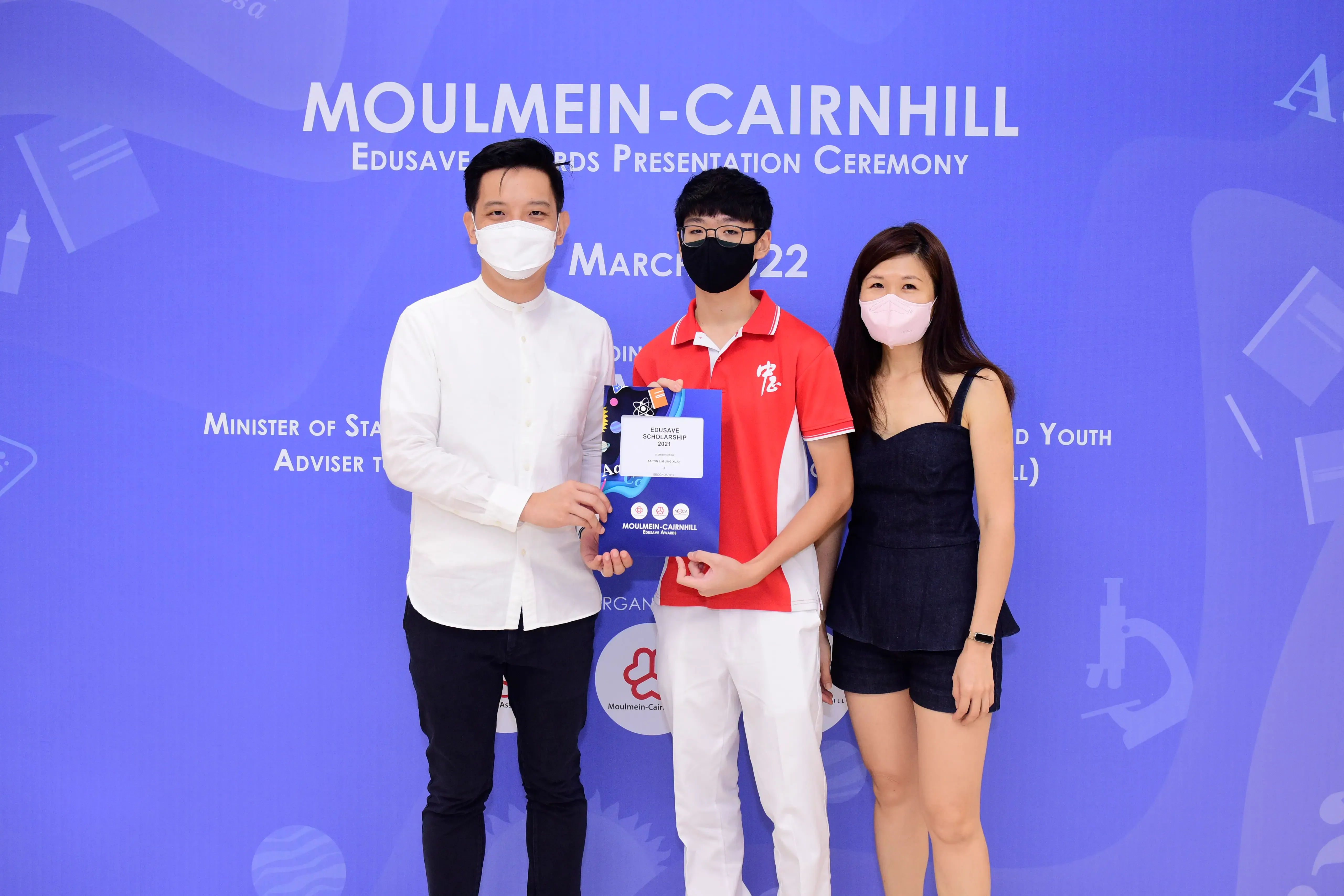Three masked people pose at the Moulmein-Cairnhill Edusave Awards Presentation Ceremony, with a student holding a certificate.