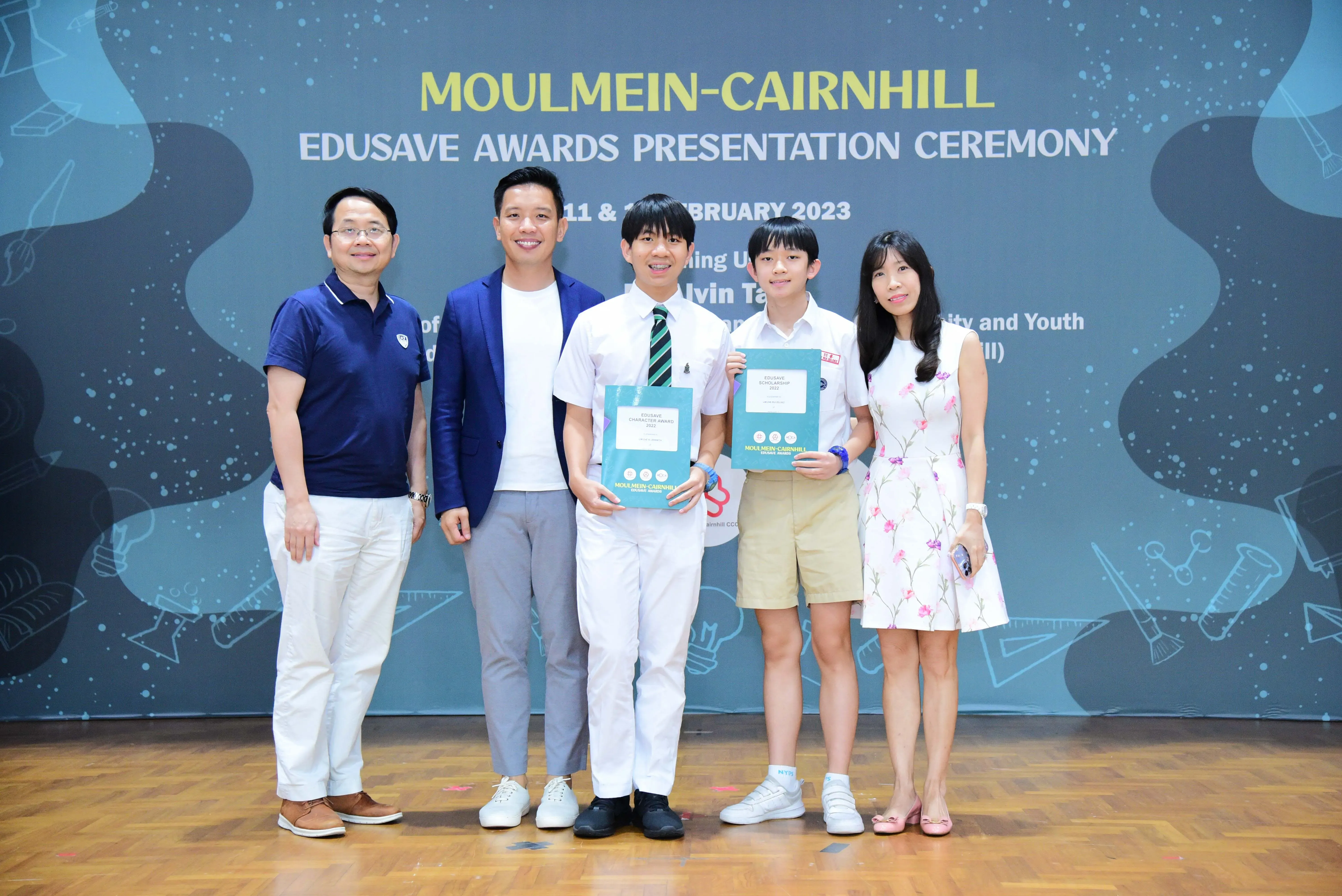 Five people pose at the Moulmein-Cairnhill Edusave Awards Presentation Ceremony, two students holding teal award folders.