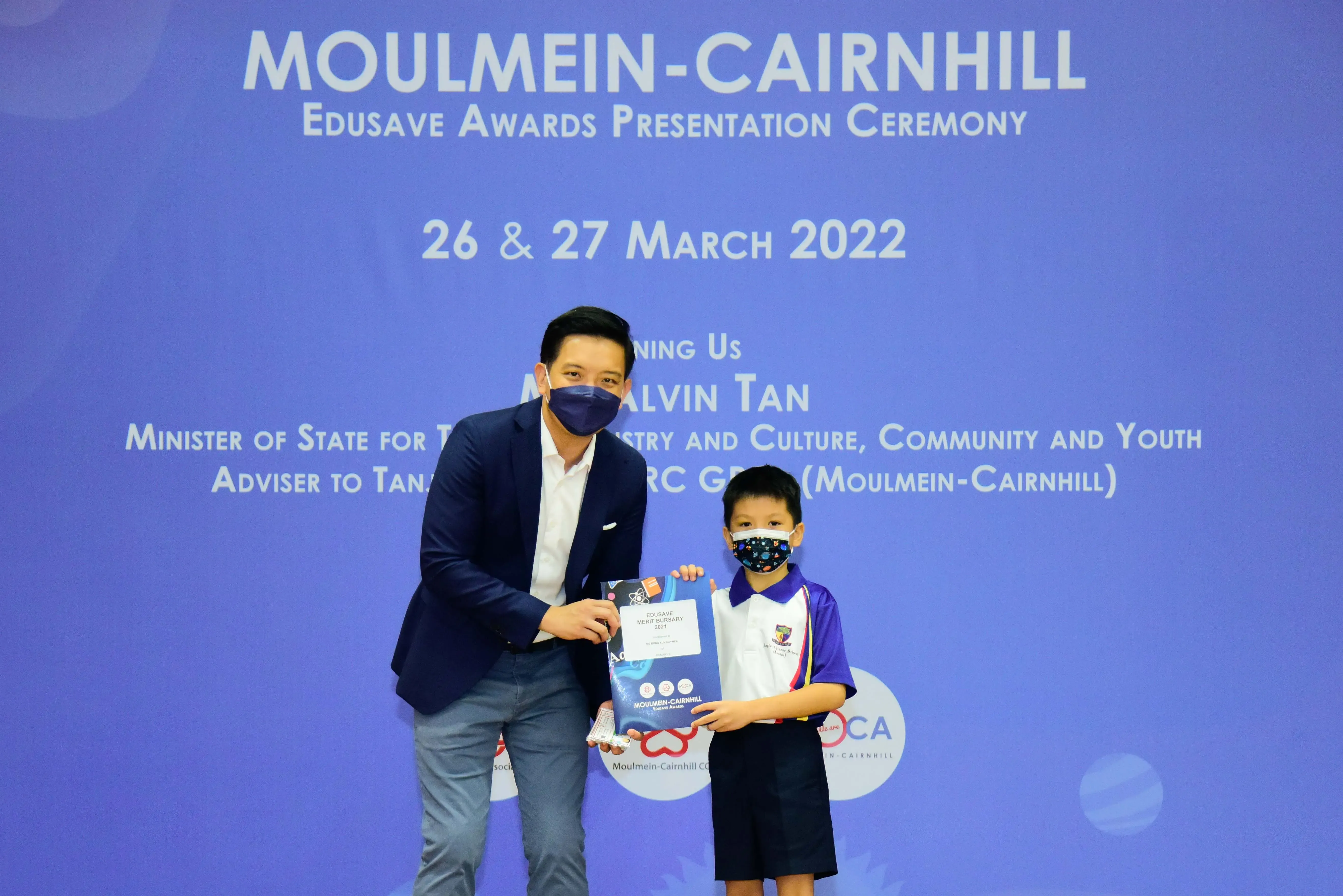An adult in a suit and a young student in uniform hold an award at the Moulmein-Cairnhill Edusave Awards Ceremony 2022.