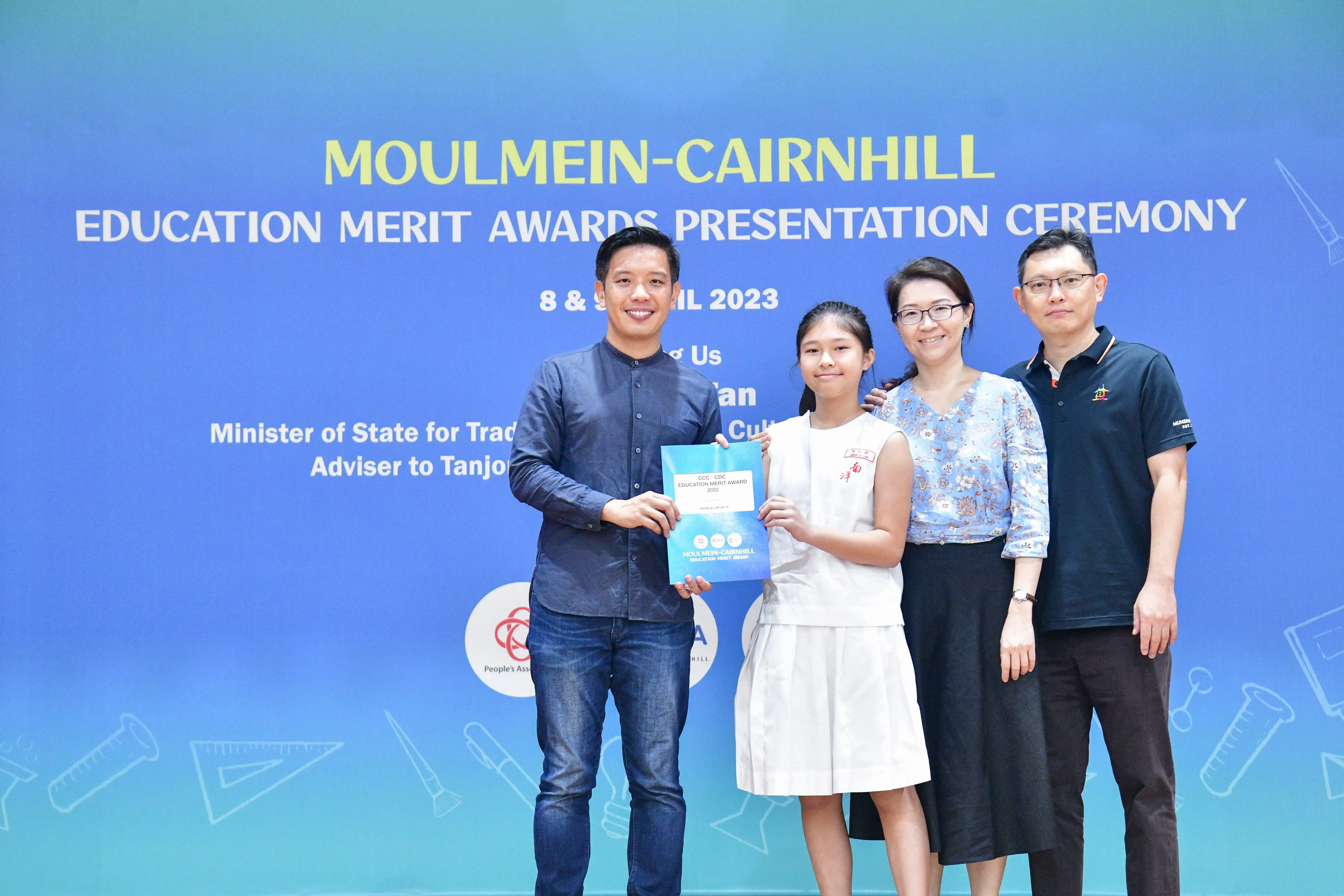 Four people pose at the Moulmein-Cairnhill Education Merit Awards Presentation Ceremony, with a girl holding a certificate.