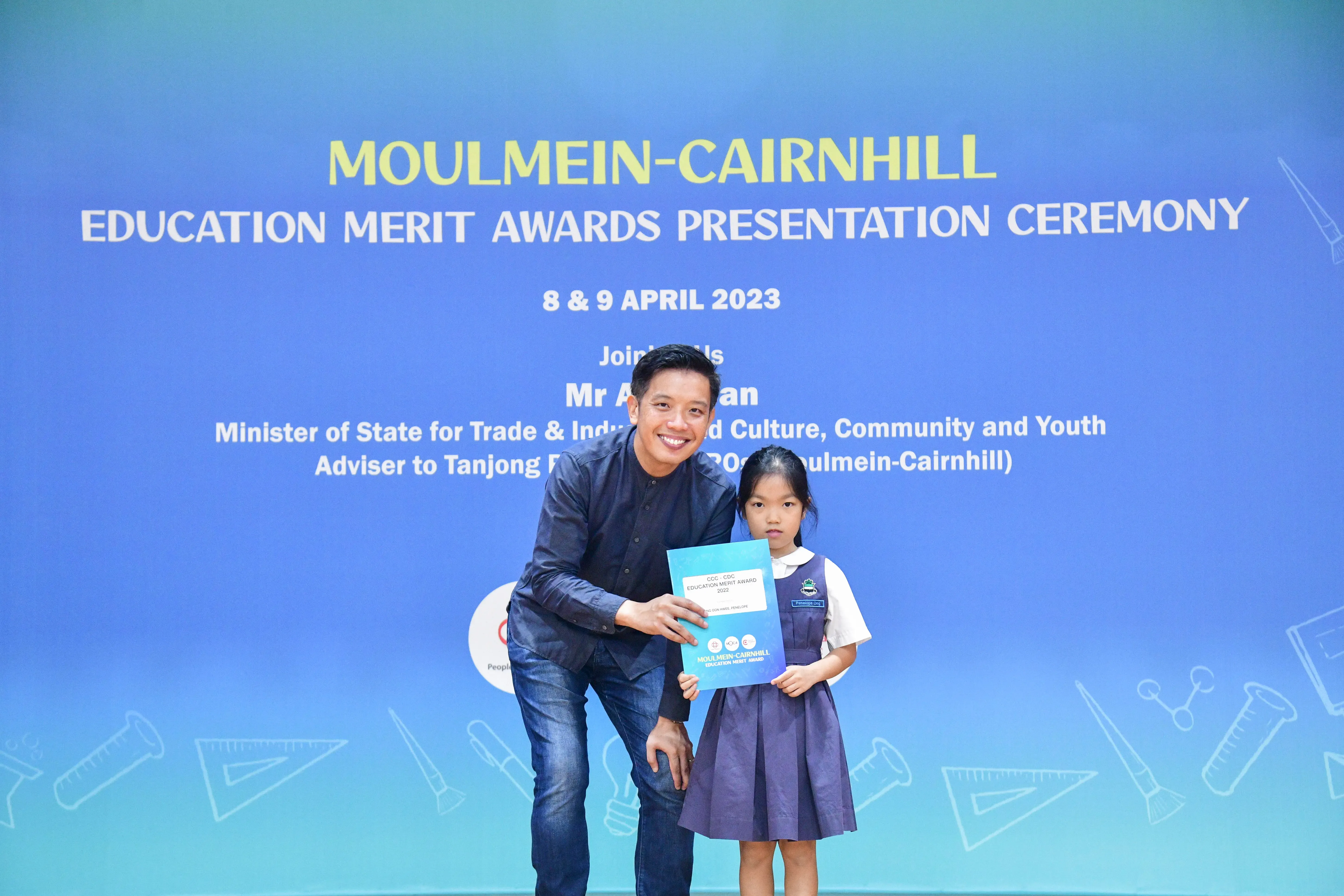 A man and a young girl hold a certificate in front of a Moulmein-Cairnhill Education Merit Awards banner dated April 2023.