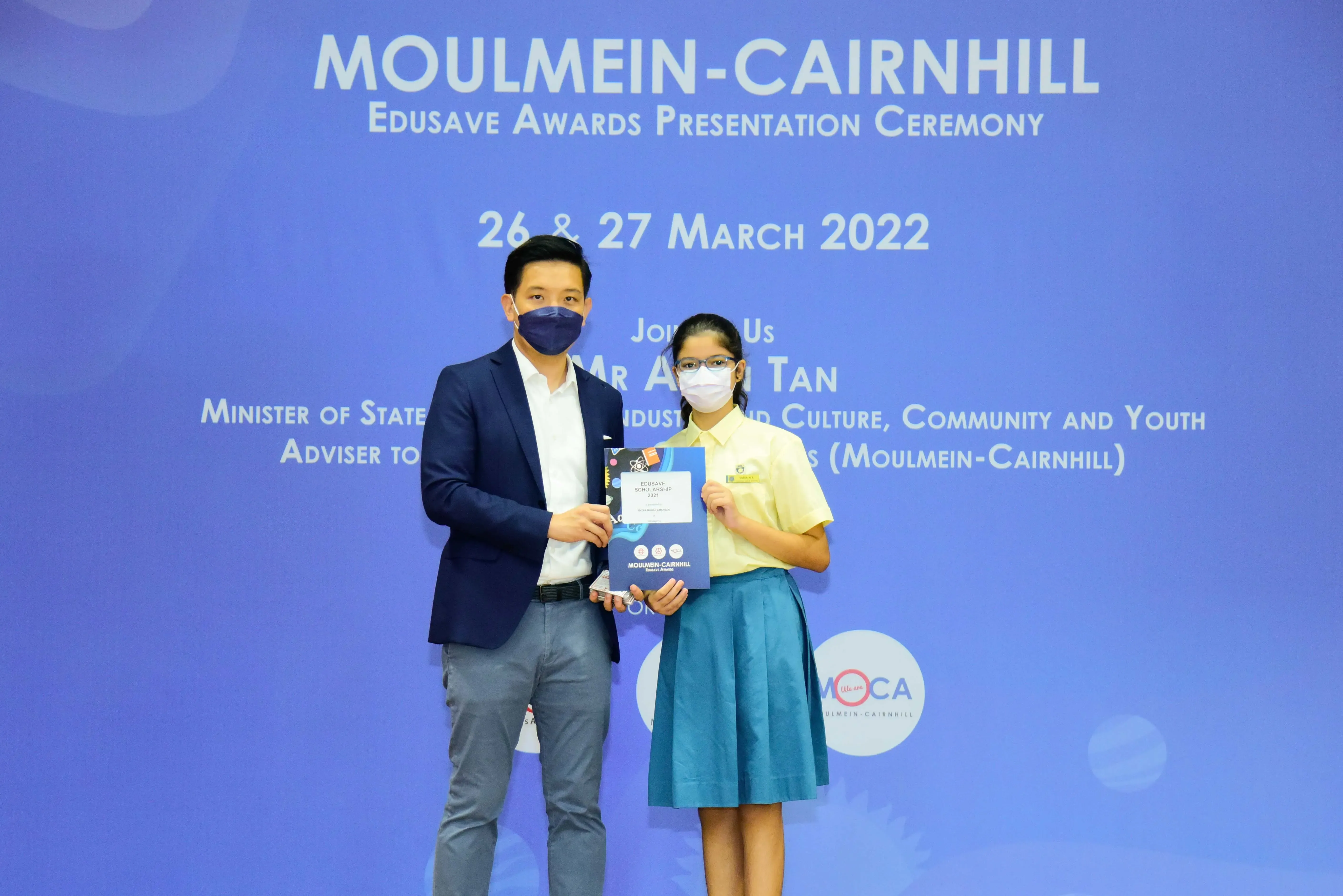 Two masked individuals hold an Edusave Scholarship folder at the Moulmein-Cairnhill Awards Ceremony, March 2022.