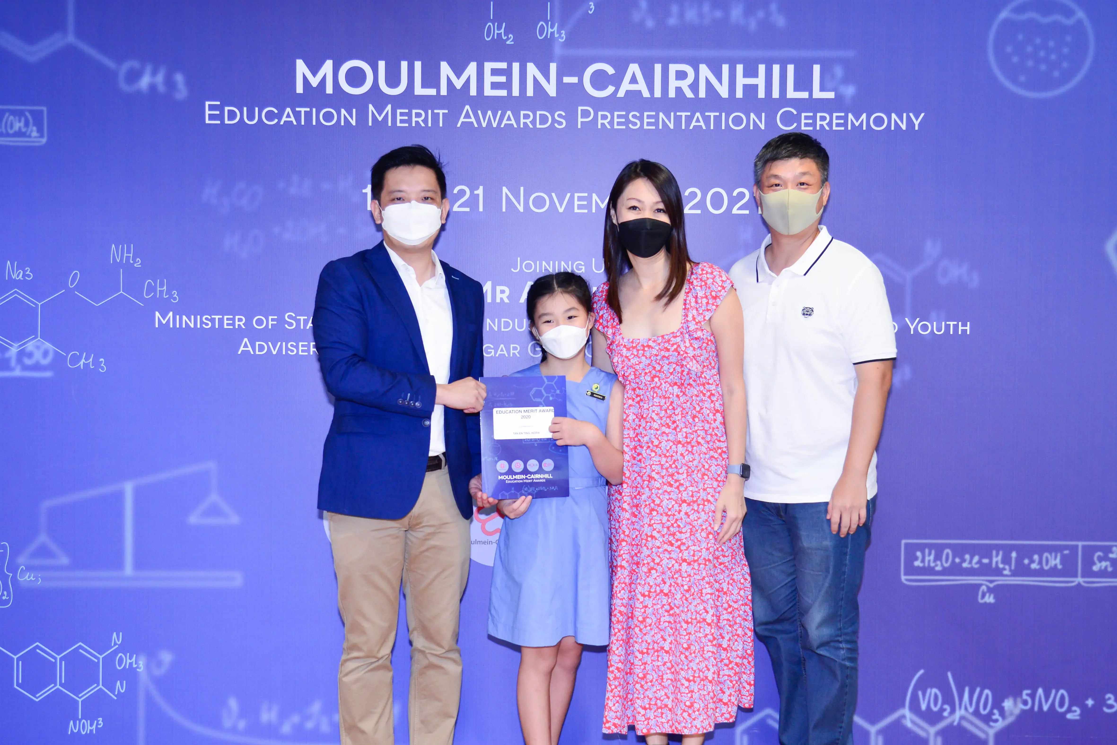 Four masked people pose at the Moulmein-Cairnhill Education Merit Awards ceremony, with a girl holding an award booklet.