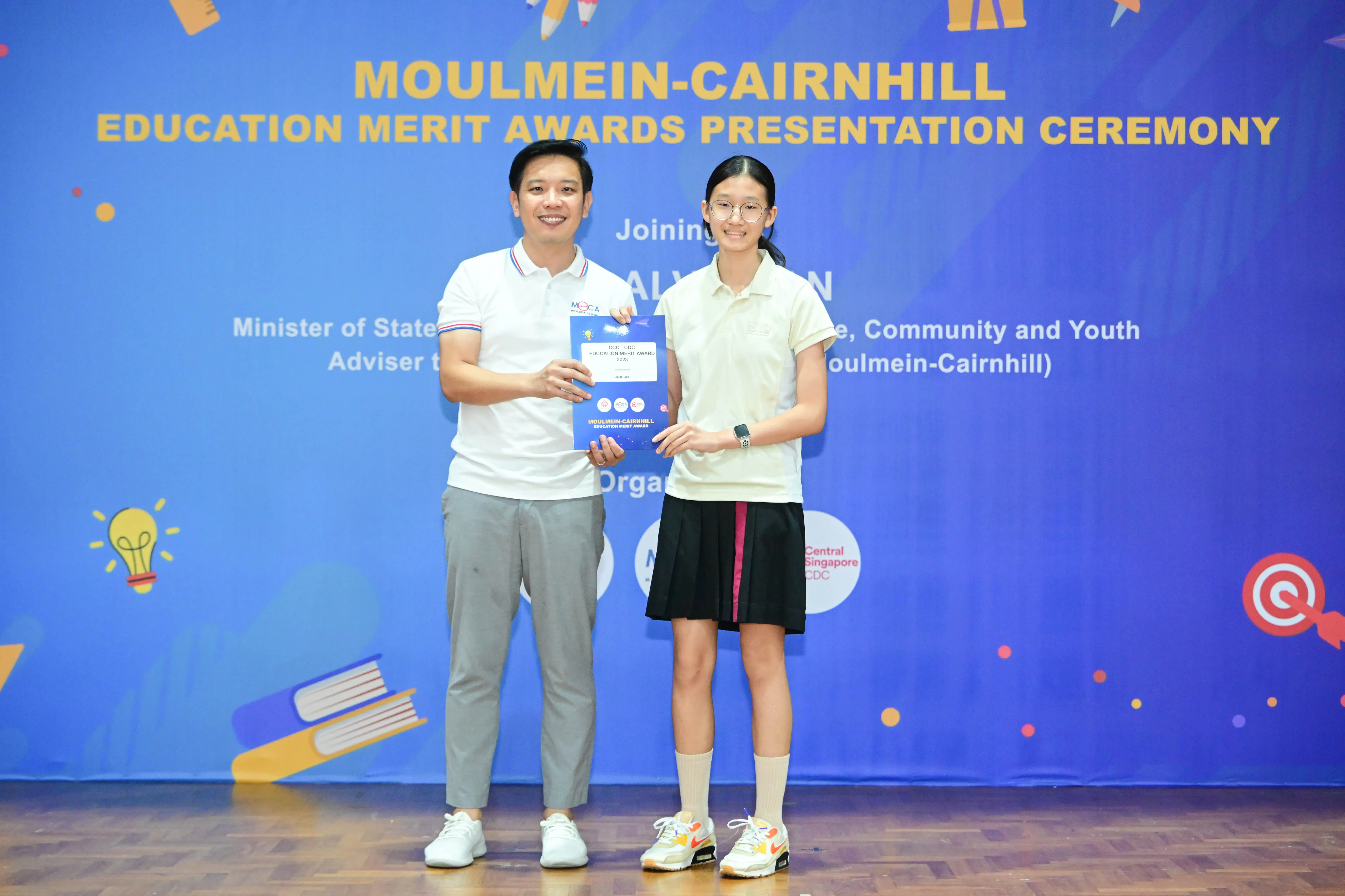 Two people holding an award certificate in front of a Moulmein-Cairnhill Education Merit Awards Presentation Ceremony banner.