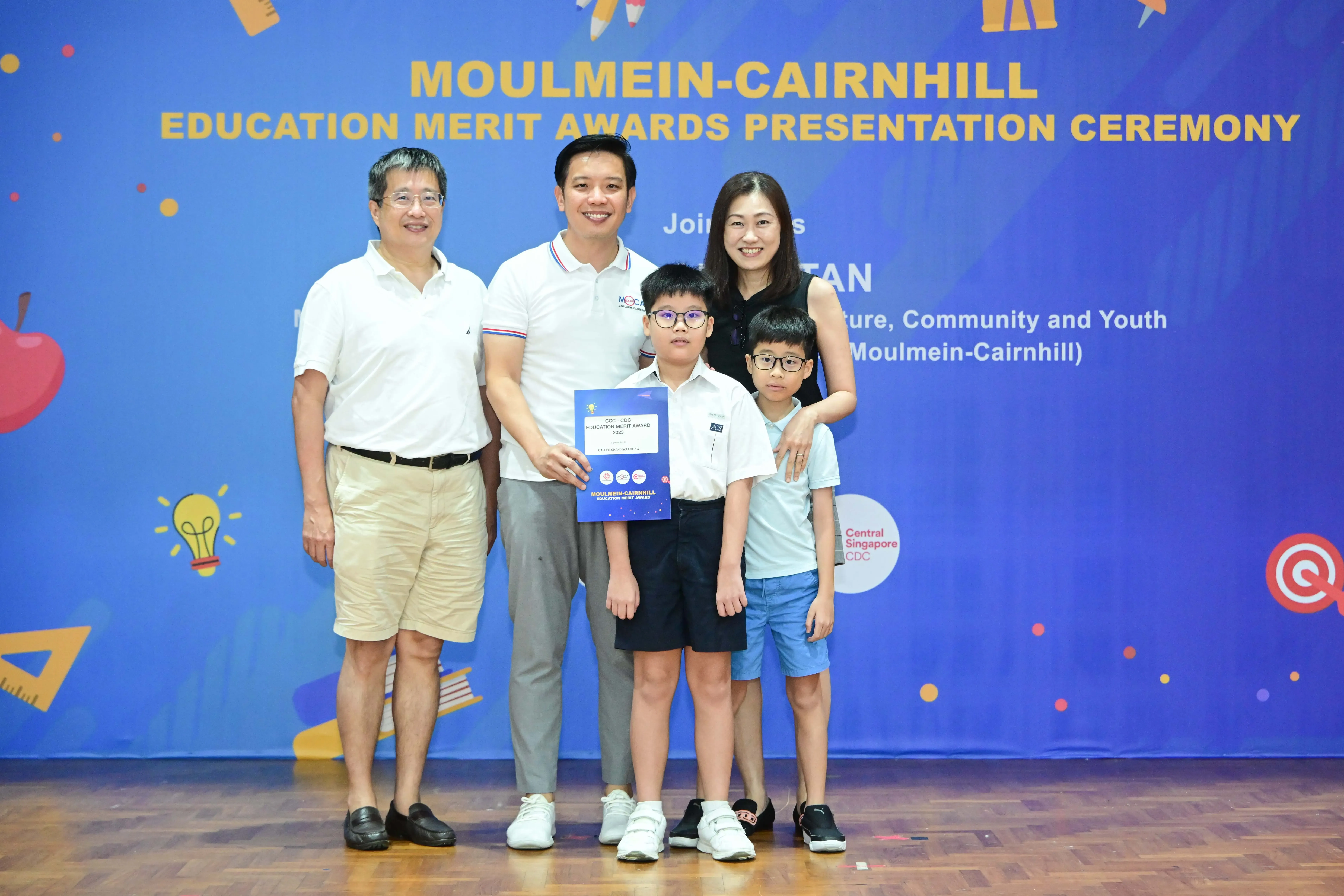 Five people pose before a Moulmein-Cairnhill Education Merit Awards banner, including two children, one holding a certificate.