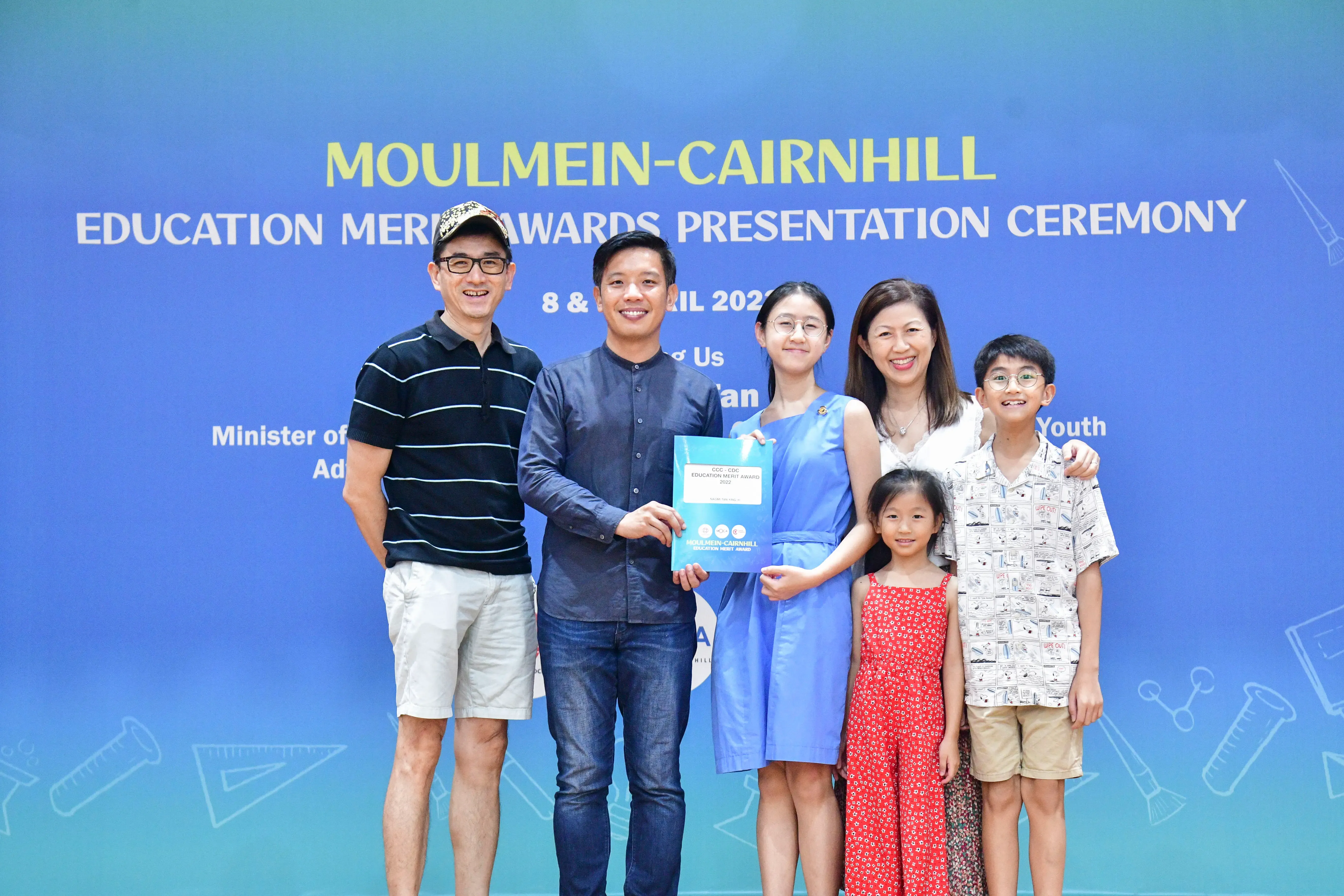 A family of five poses with a man holding a certificate at the Moulmein-Cairnhill Education Merit Awards ceremony.
