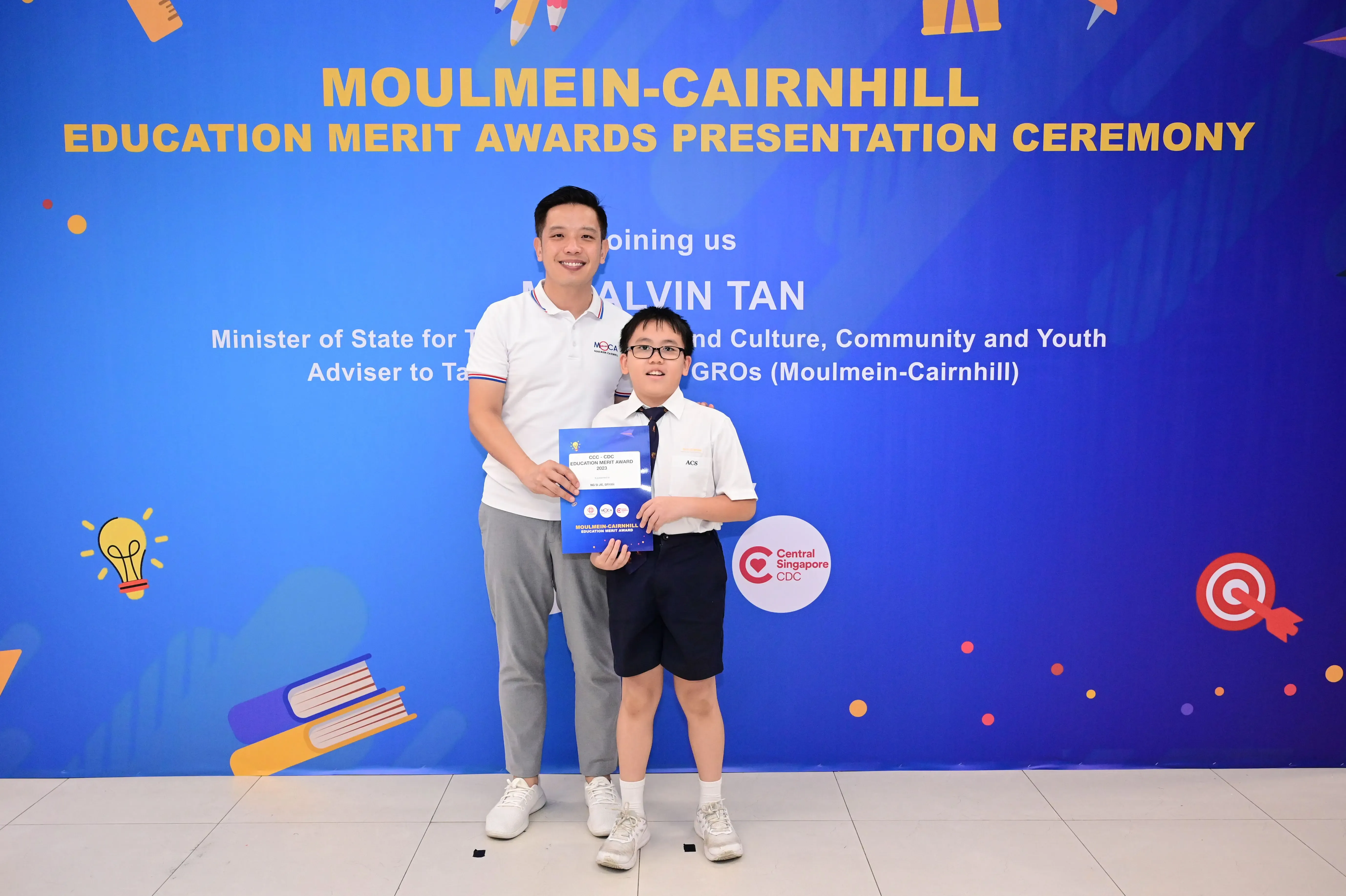 A man and a young boy hold an award certificate in front of a Moulmein-Cairnhill Education Merit Awards banner.