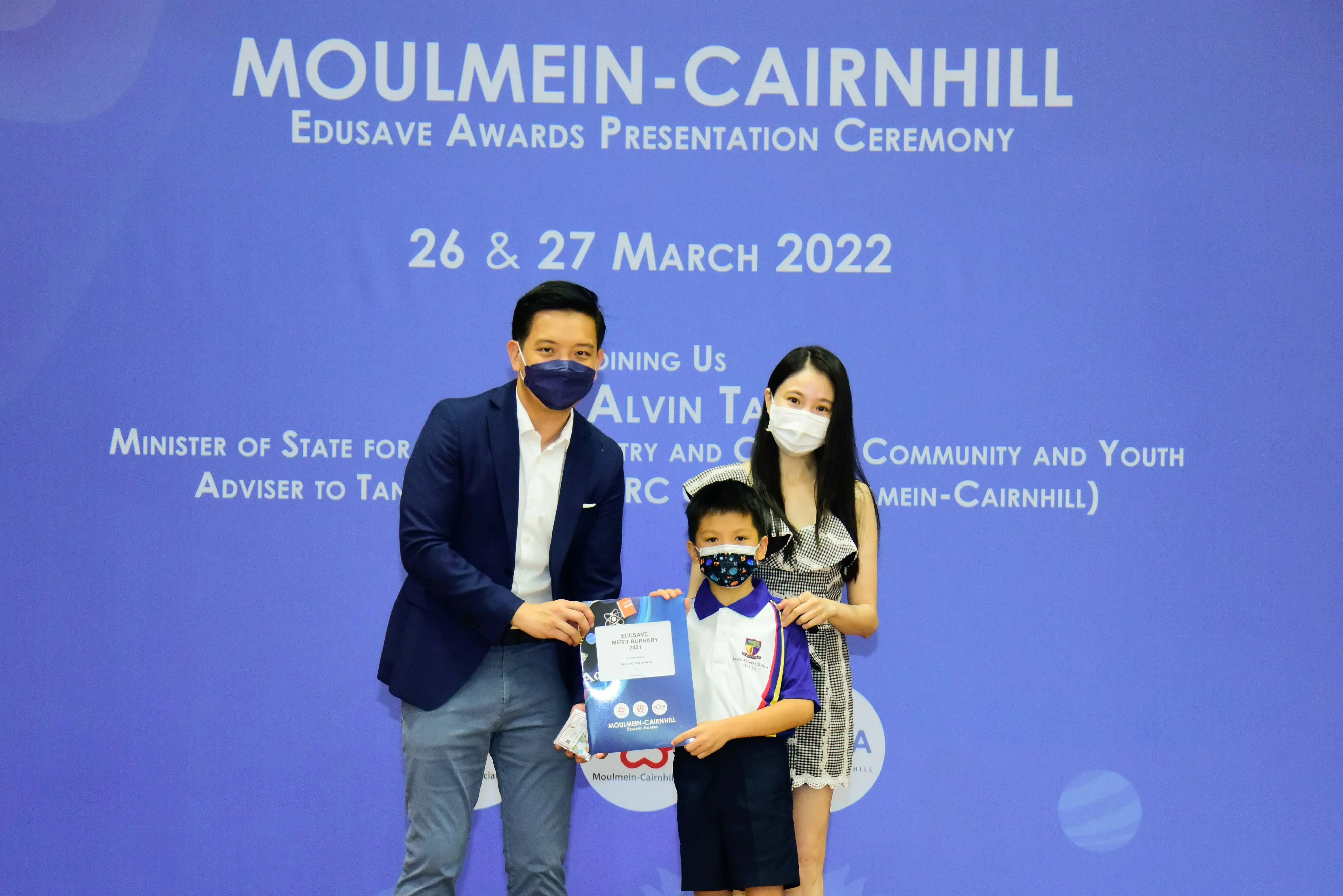 Two adults and a young child in masks hold an award at the Moulmein-Cairnhill Edusave Awards Ceremony, March 2022.