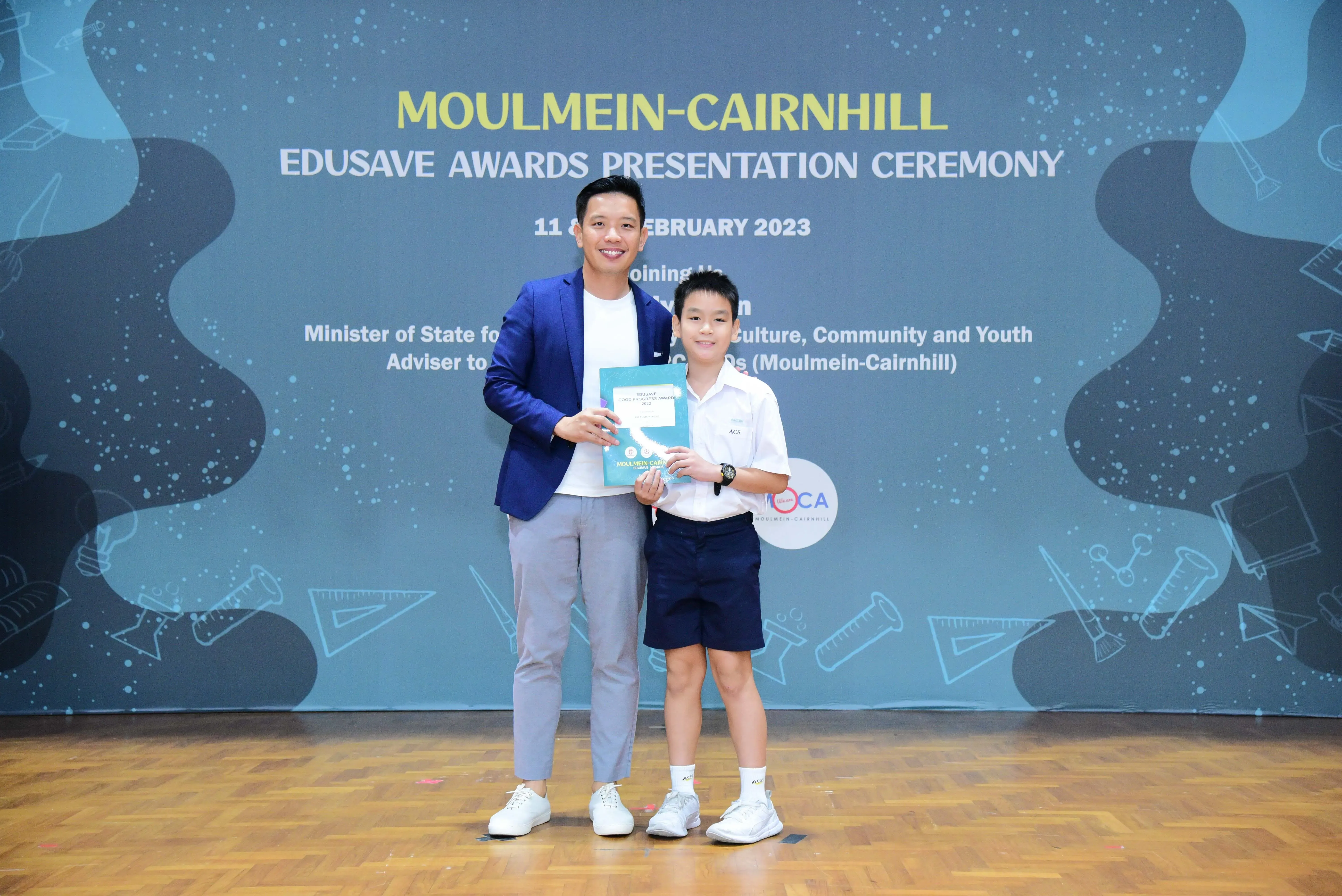 A man in a blue blazer and a young boy in school uniform pose holding certificates at the Moulmein-Cairnhill Edusave Awards 2023.