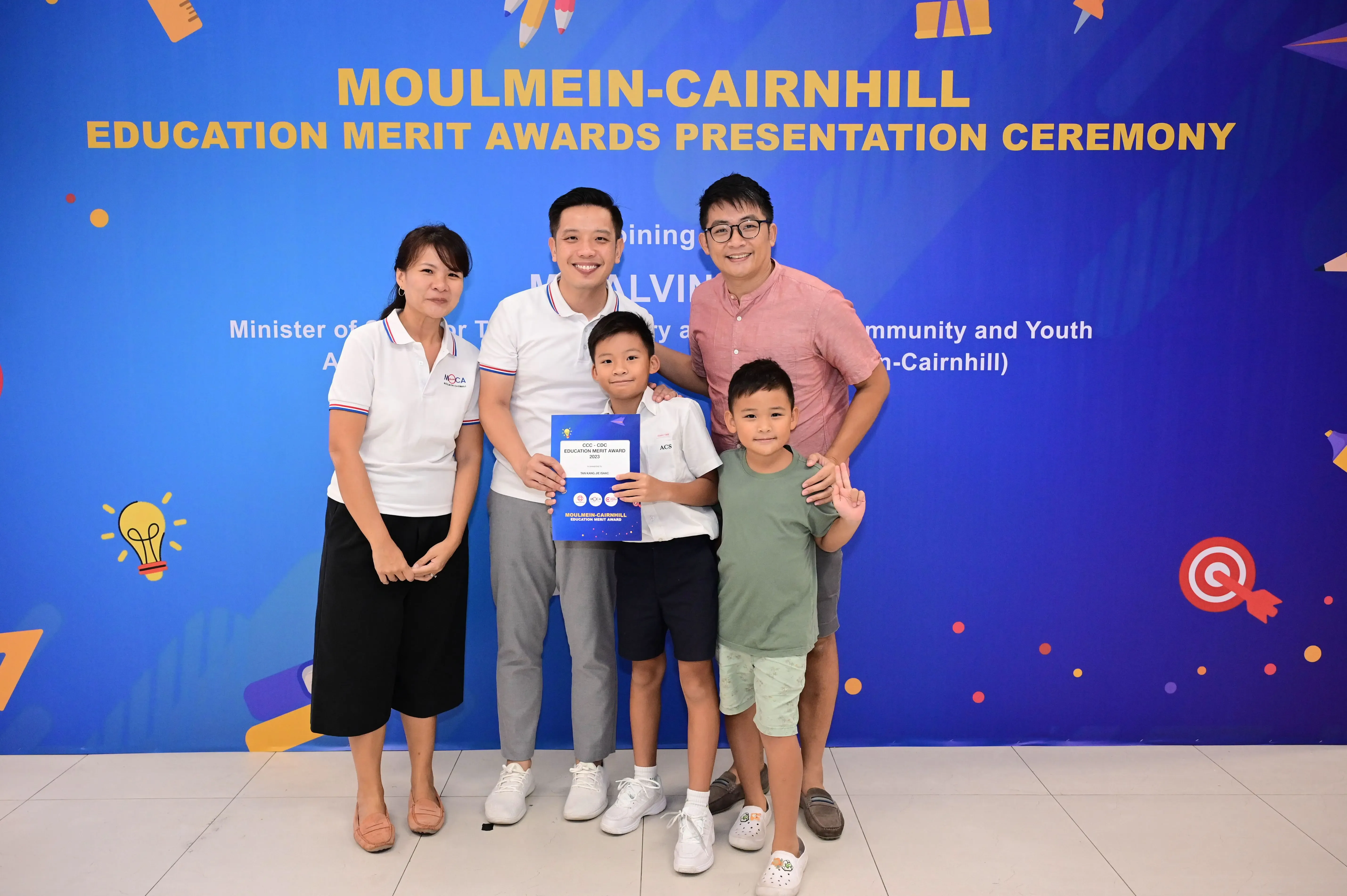 Five people pose before a blue Moulmein-Cairnhill Education Merit Awards banner, with a boy holding a blue certificate.