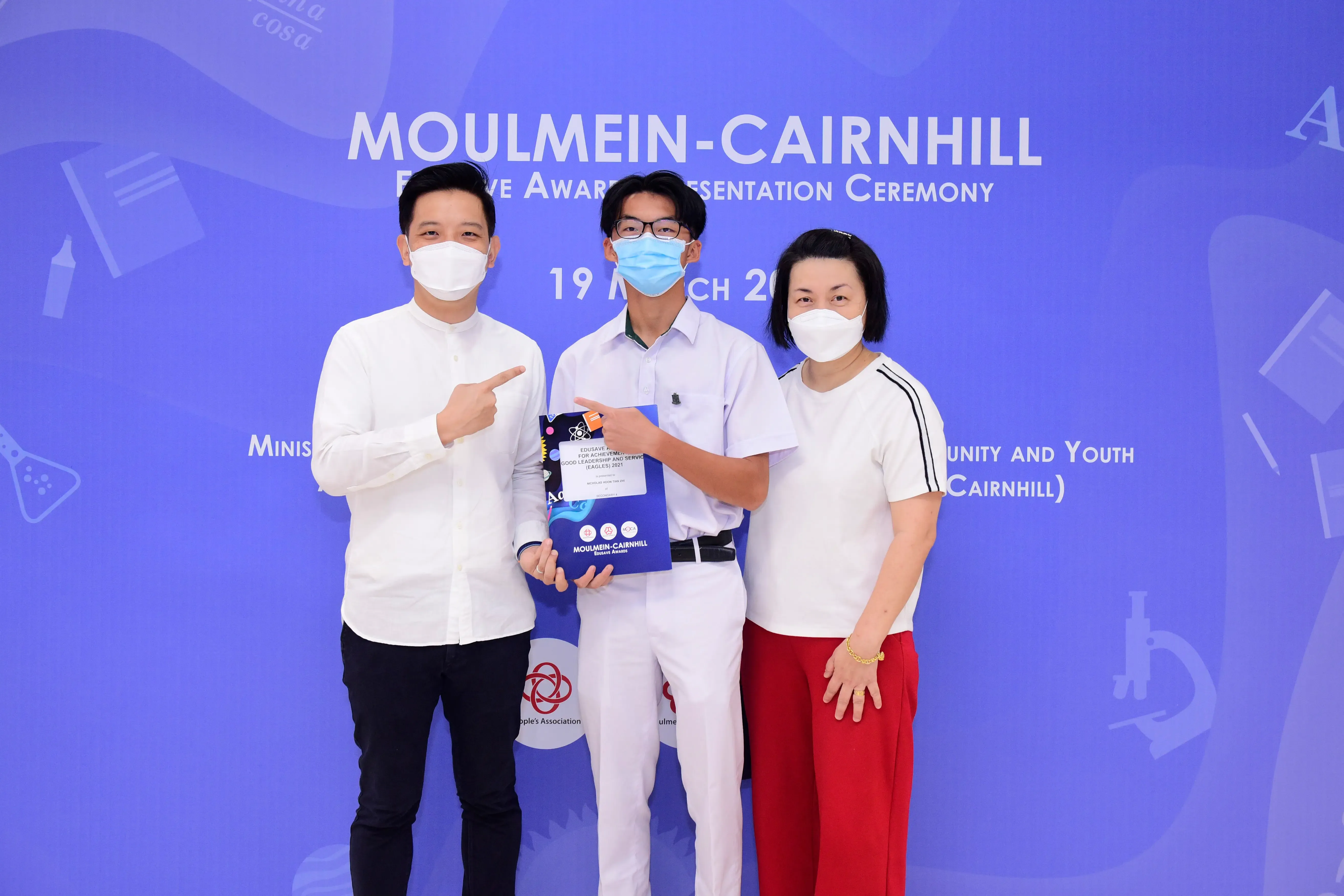 Three masked people pose at the Moulmein-Cairnhill Awards Presentation Ceremony, with the center person holding an award.