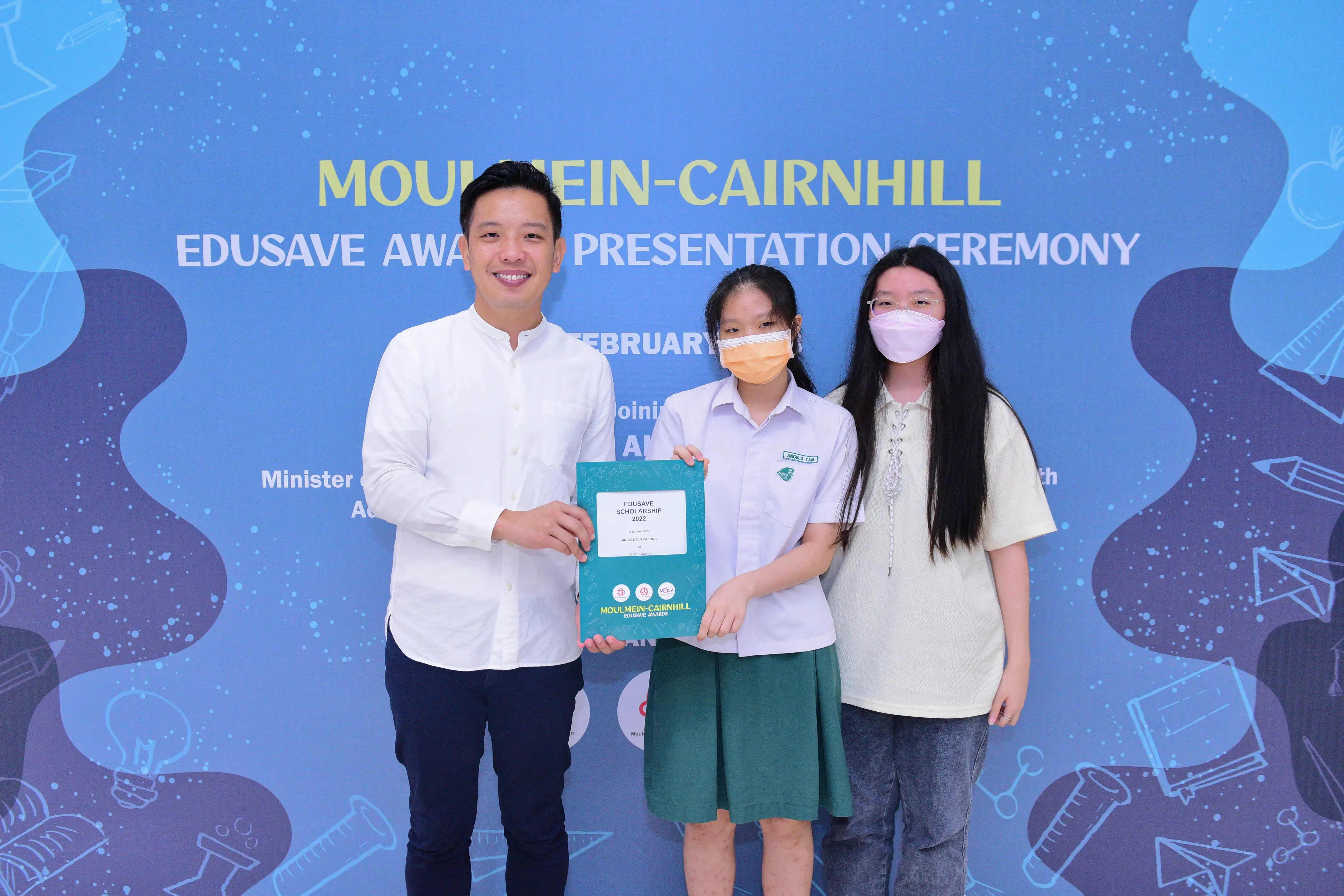 Three people pose at the Moulmein-Cairnhill Edusave Award Presentation Ceremony, holding a scholarship certificate.