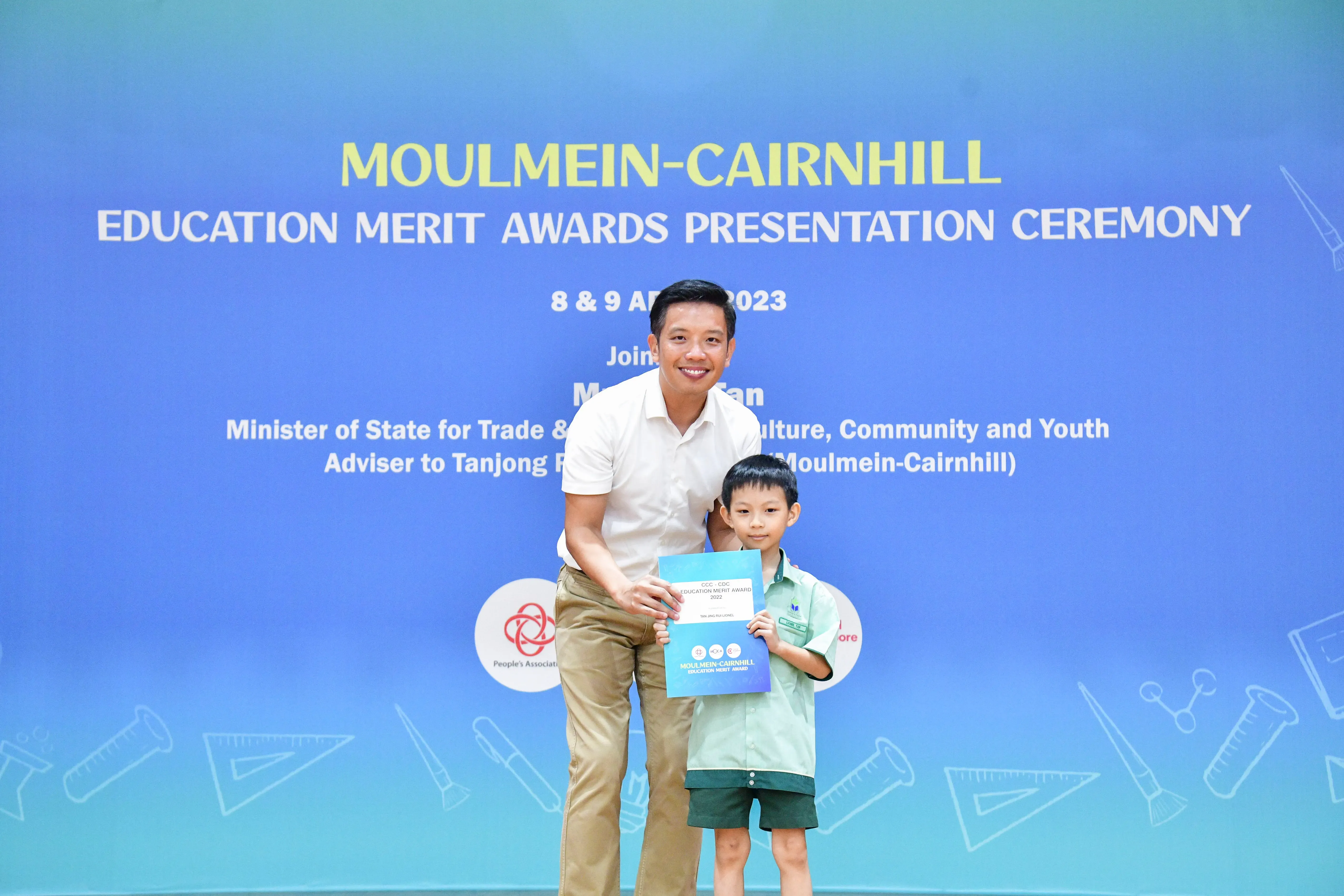 A man and a young boy holding a certificate pose in front of the Moulmein-Cairnhill Education Merit Awards banner.